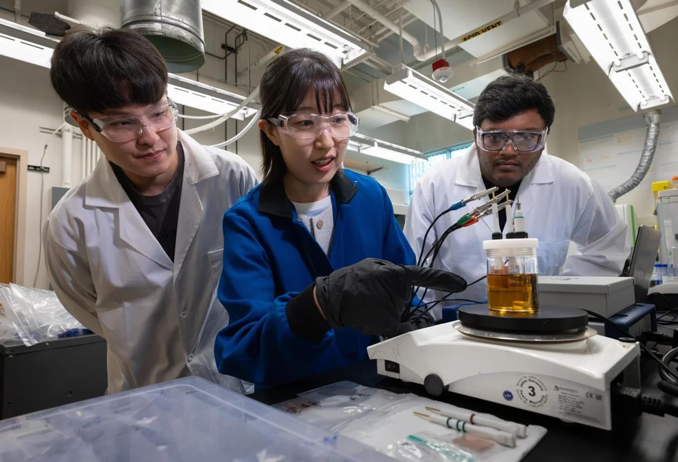 Three scientists in lab coats and safety glasses examine a jar with electrodes on a rotating platform in a lab.