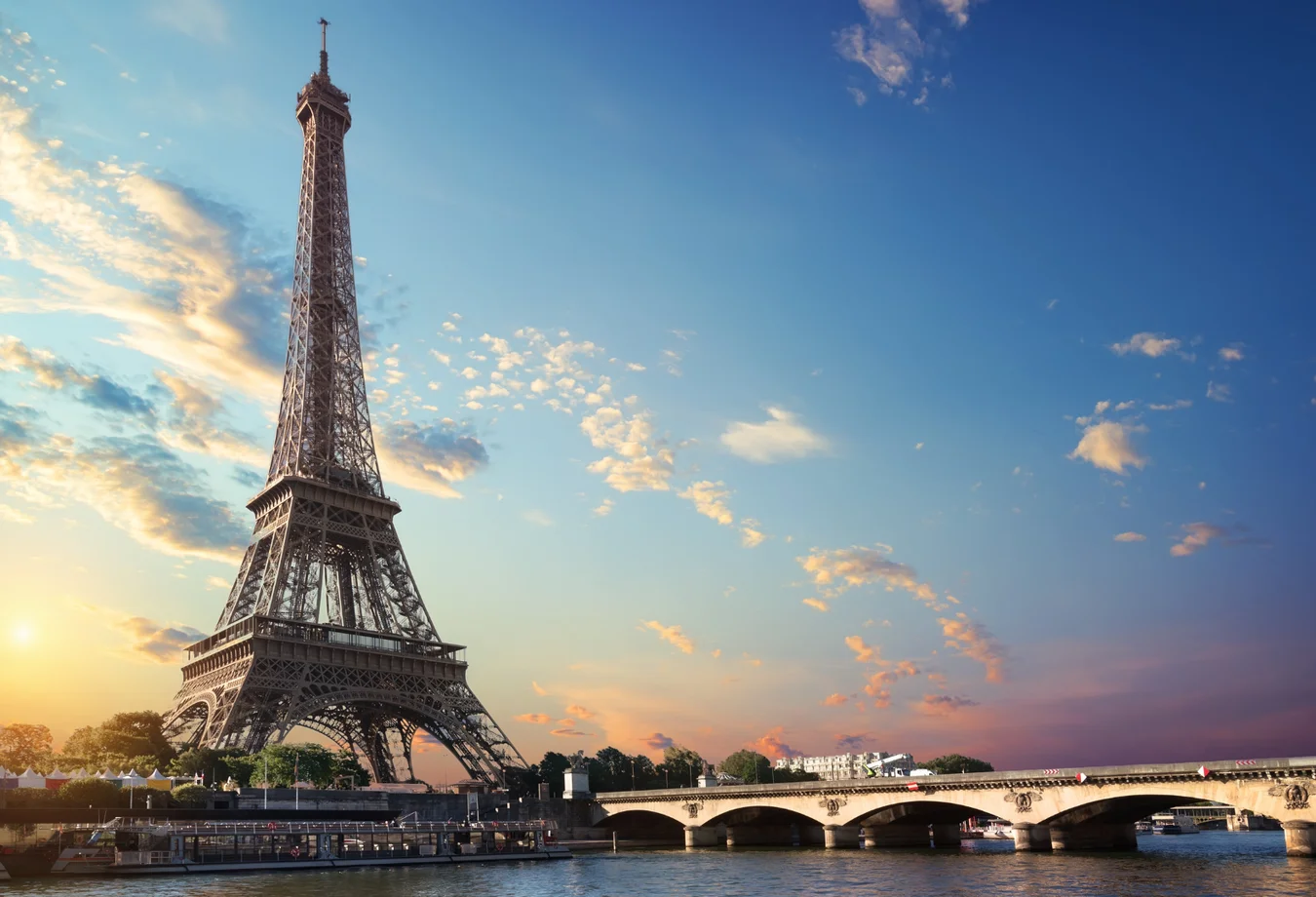 Eiffel Tower at sunset with partly cloudy sky and a bridge over the Seine River in Paris.