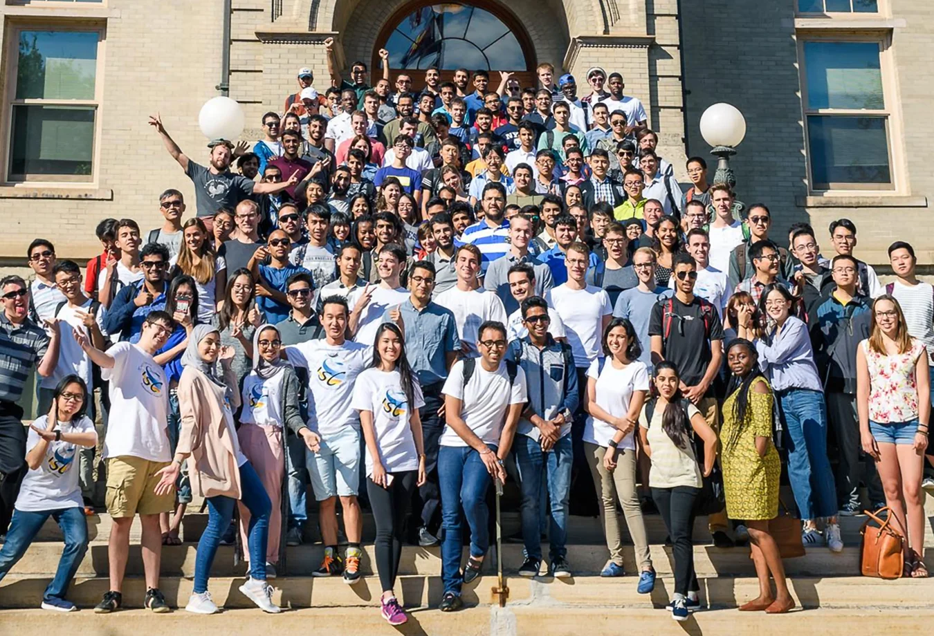 Large group of international students posing on outdoor steps in front of a building, some wearing matching white shirts.
