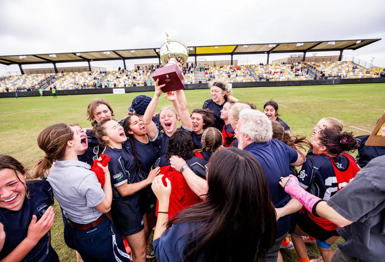 Women's rugby team celebrating victory and holding a large trophy on a grassy field with spectators in the stands behind them.