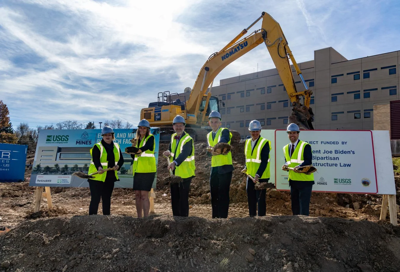Six people in safety vests and helmets hold shovels during a groundbreaking ceremony at the USGS building construction site with an excavator and signage.
