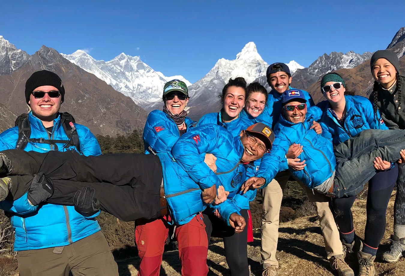 Group of students in blue jackets smiling and holding a person horizontally with snowy mountain peaks in the background.