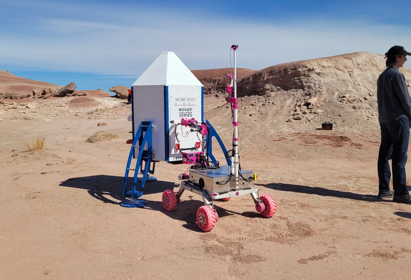 A man standing next to a small robot in the desert.
