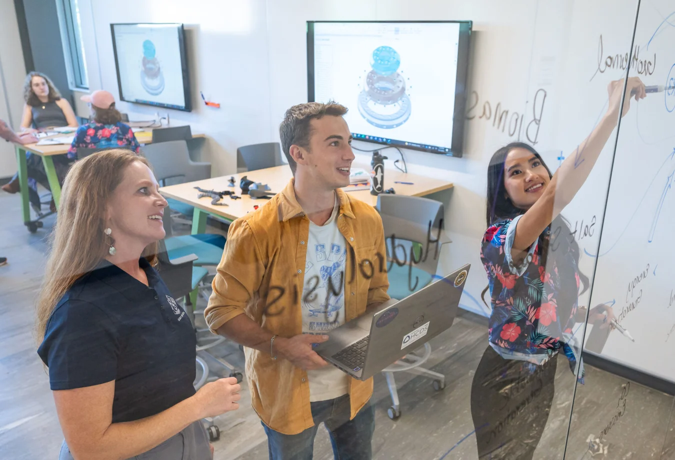 Engineering Design students and professor collaborating in a modern office, brainstorming ideas on a glass board with laptops and digital tech in a bright workspace.