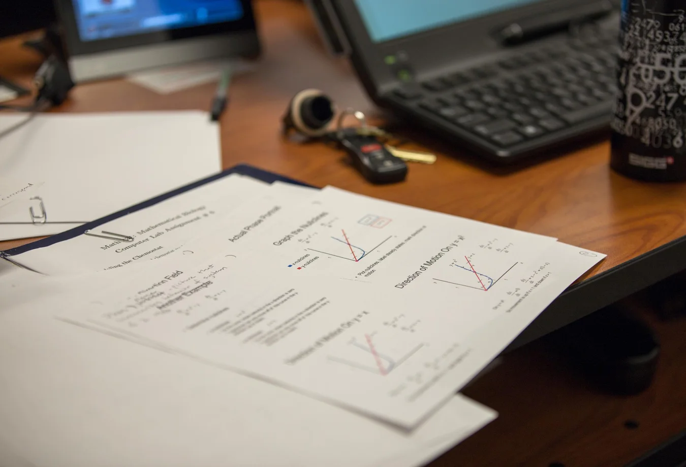 Desk with math notes featuring graphs, a laptop keyboard, car keys, and a black patterned water bottle.