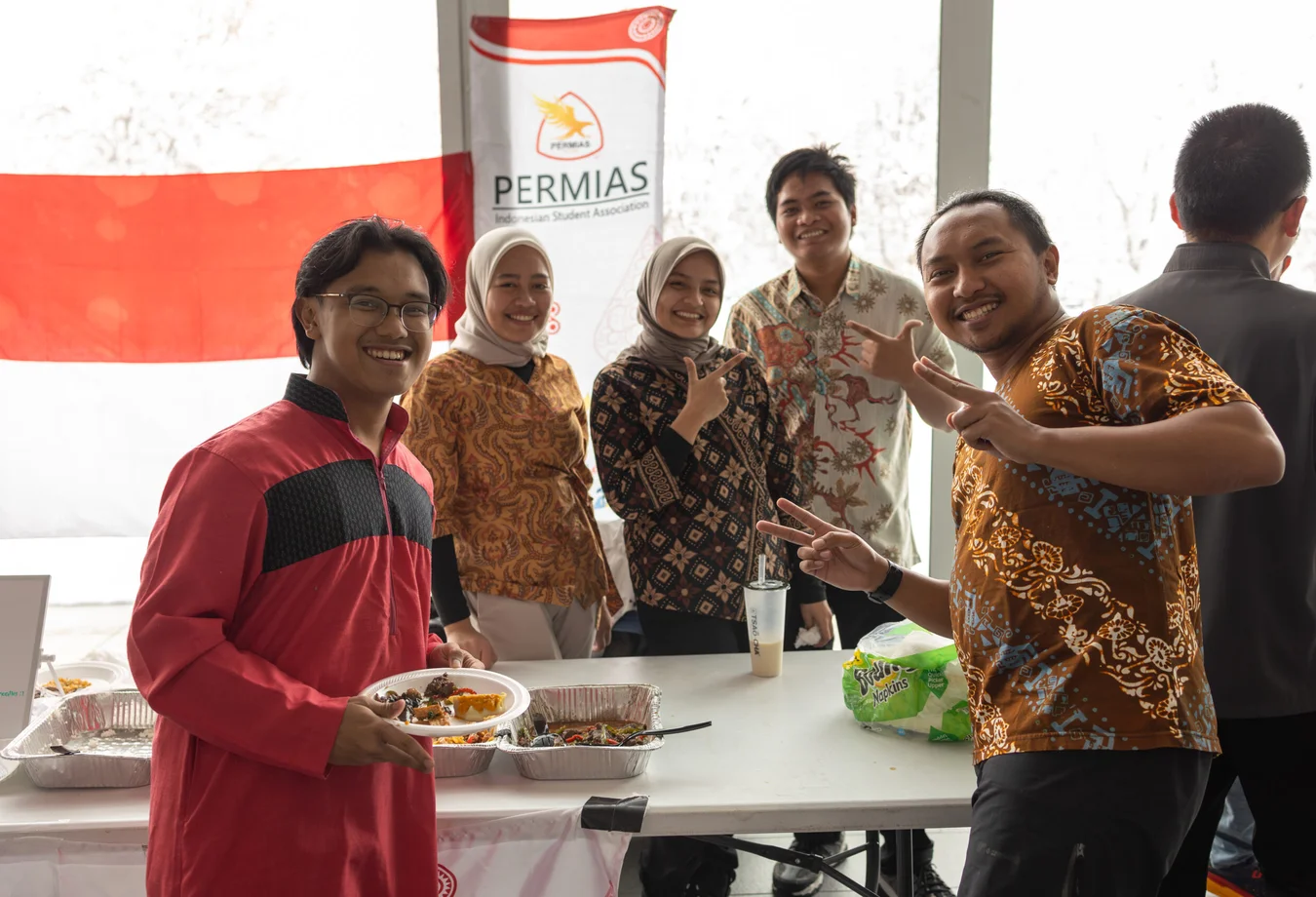 Group of smiling students in batik and traditional clothing posing at a PERMIAS booth with food trays and Indonesian flag.
