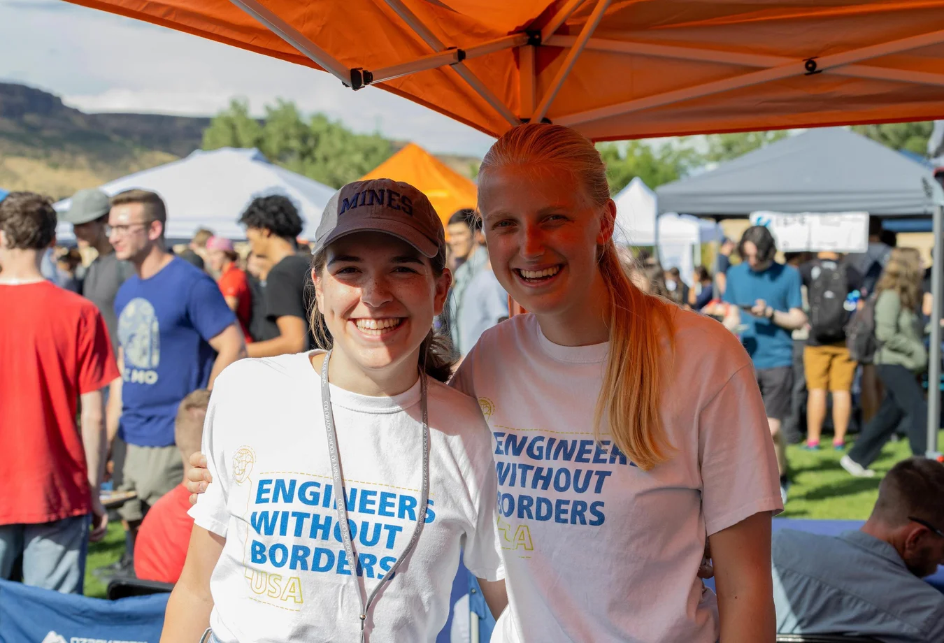 Two smiling women in “Engineers Without Borders” shirts pose under an orange canopy at a busy outdoor event.