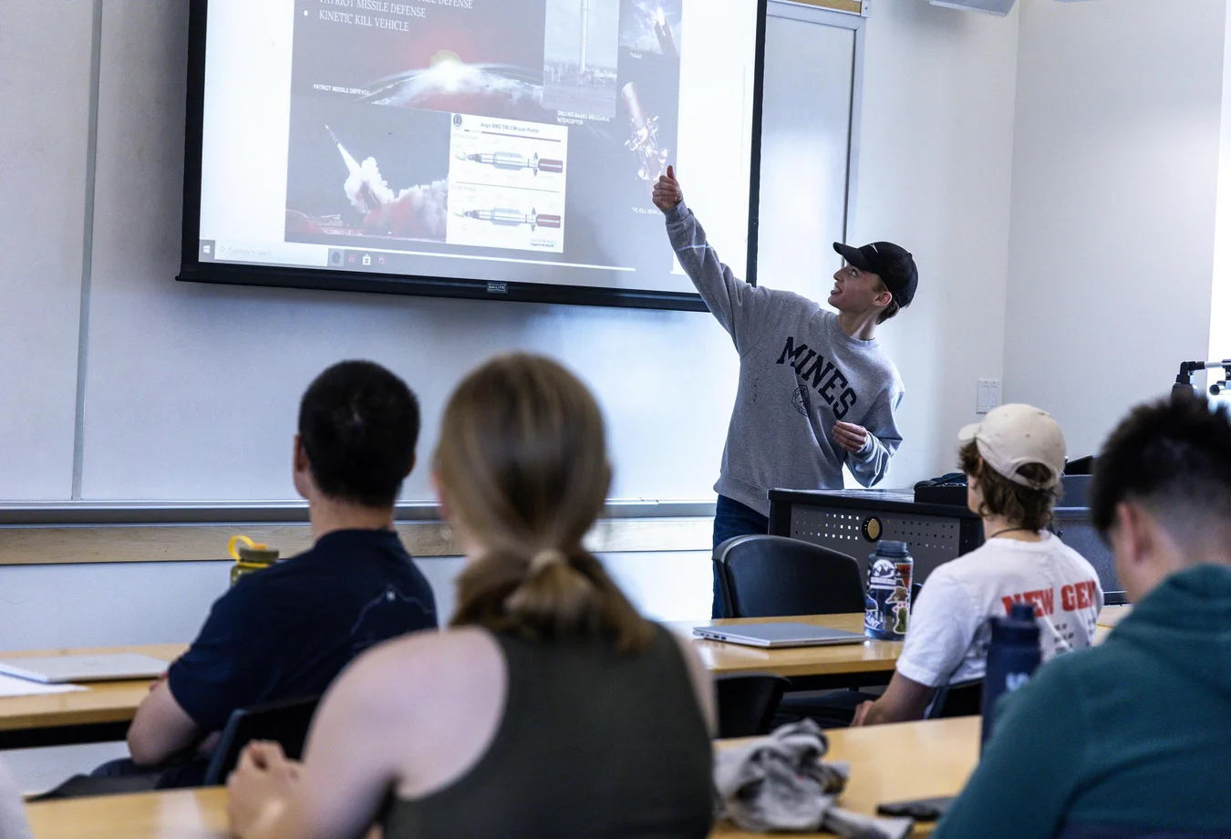 A student stands at the front of a classroom, presenting to an engaged audience of students seated at desks.