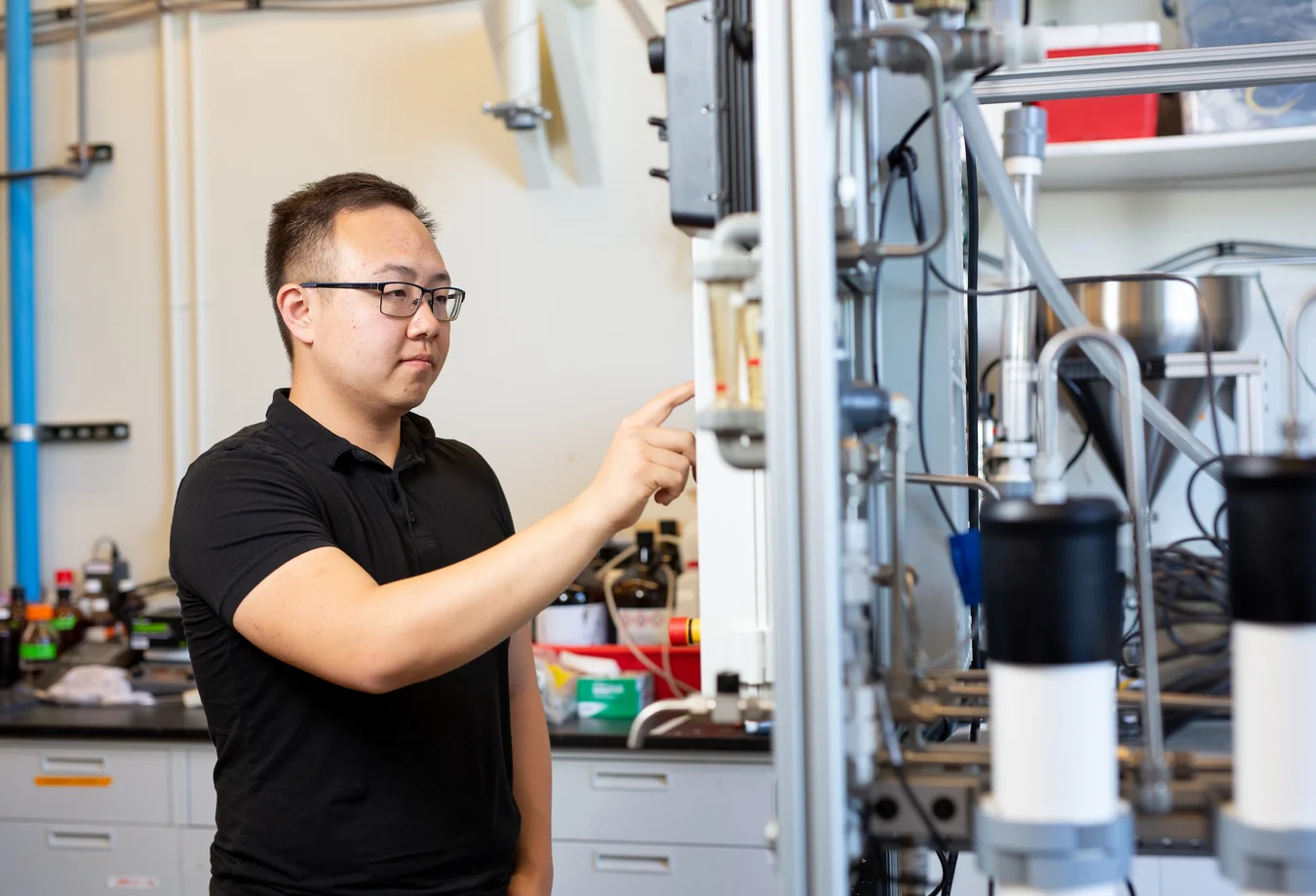 A researcher points to a transparent tube on a large laboratory testing system surrounded by tubing, pumps and other equipment.