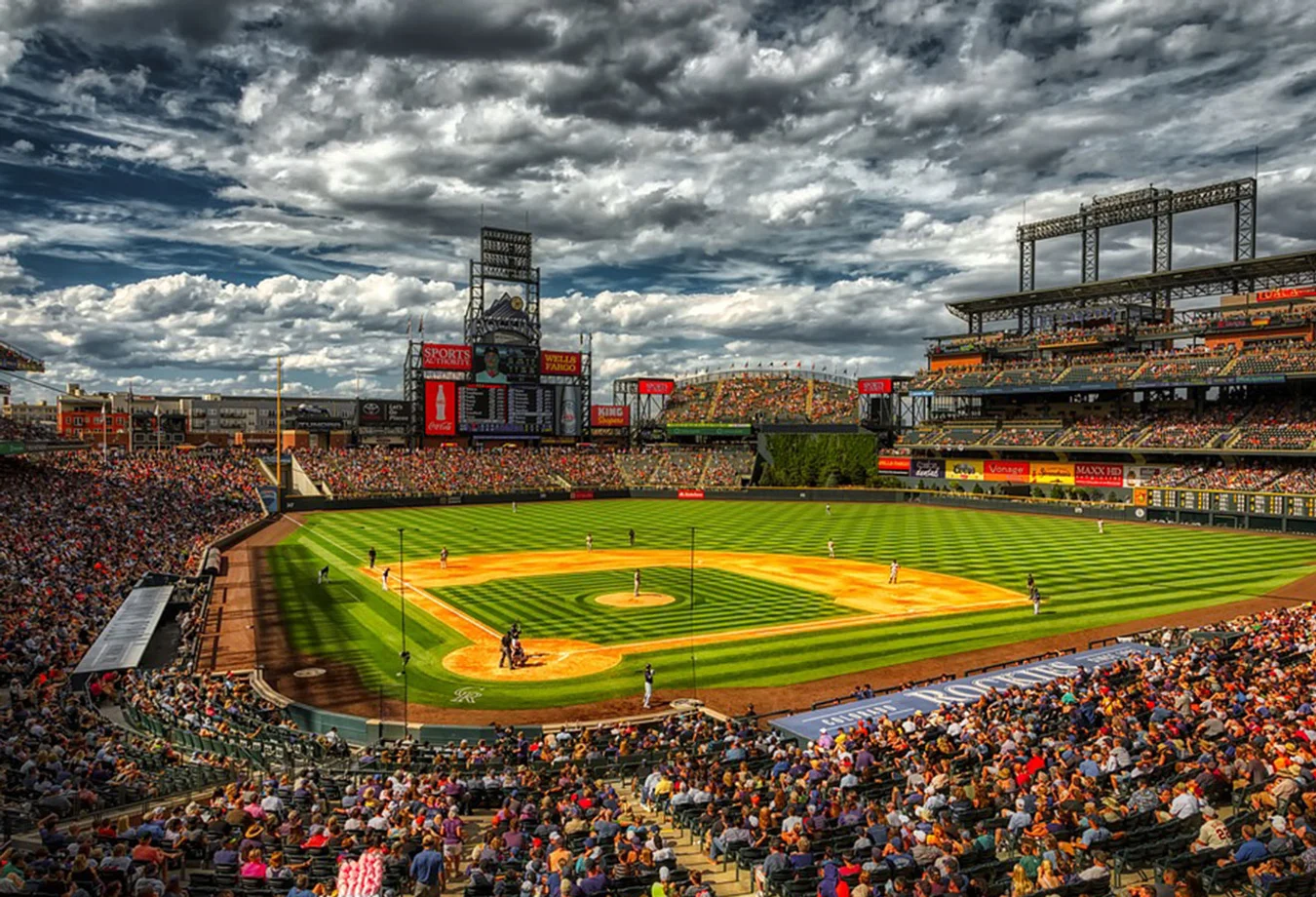 Coors Field baseball stadium under a dramatic cloudy sky during a daytime game with players on the field and fans in the stands.