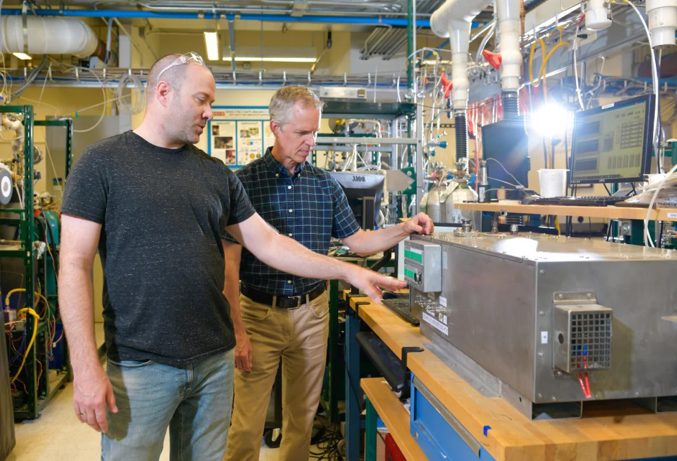 Two men examining and working with scientific equipment in a laboratory filled with instruments and computers.
