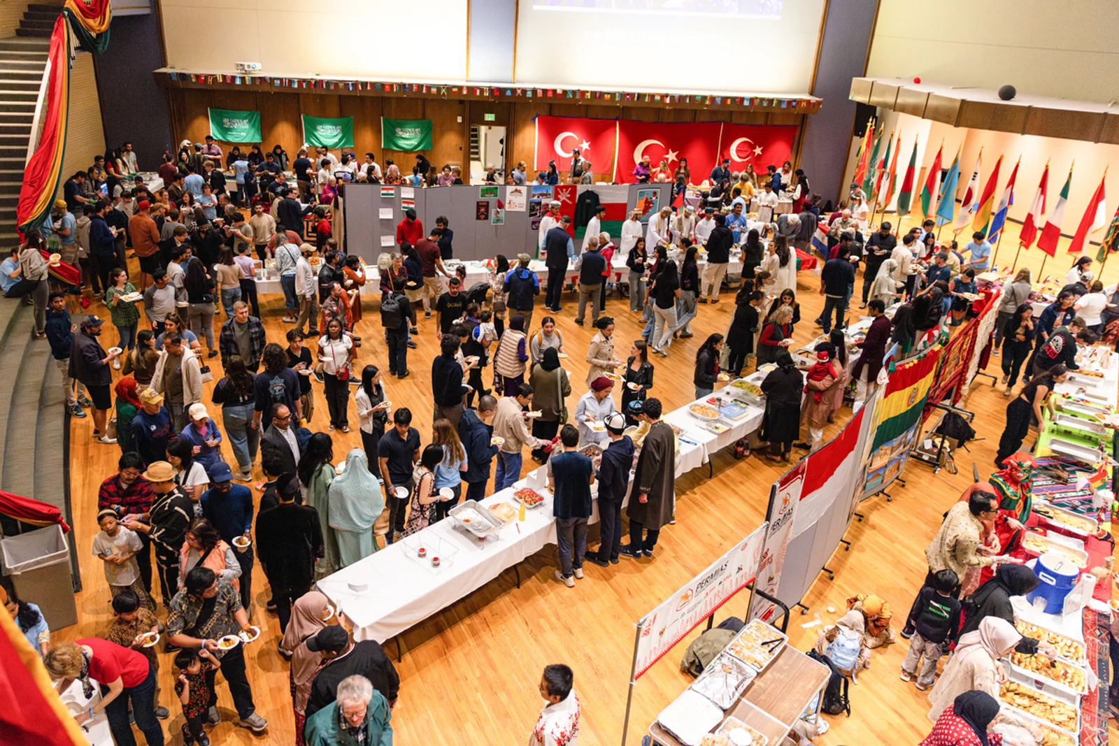 Large multicultural crowd enjoying an indoor international food festival with country flags and diverse dishes on display.