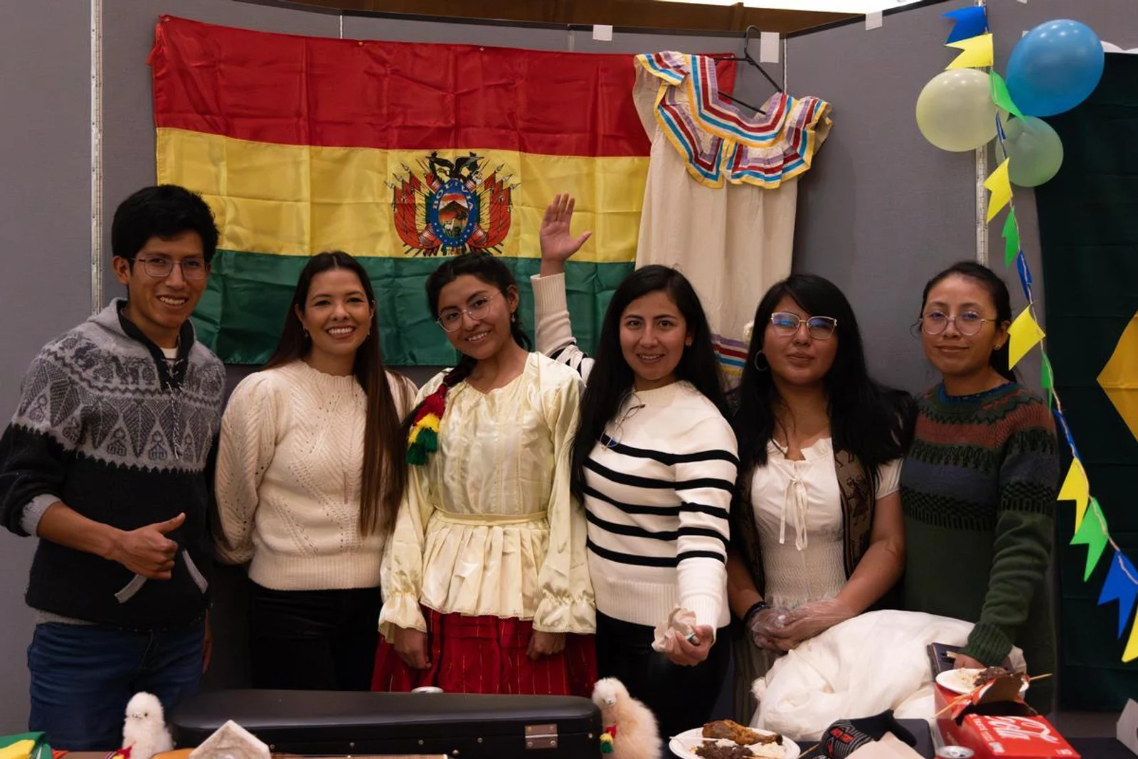 Six students smiling in front of a Bolivian flag with traditional clothing and festive decorations in the background.