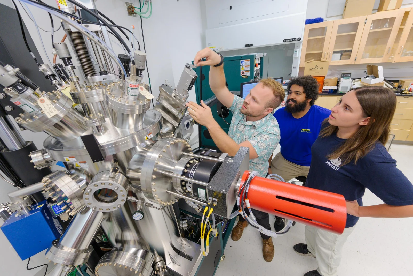 Three researchers examine a large stainless-steel vacuum apparatus in a laboratory.