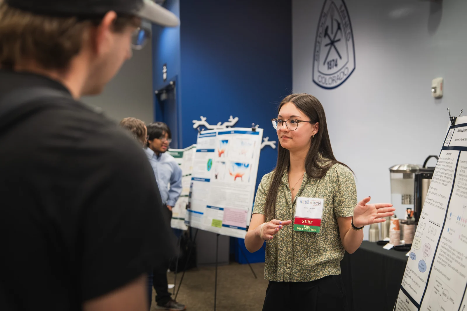Undergraduate female student wearing glasses presents a research poster to attendees at an undergraduate research symposium.