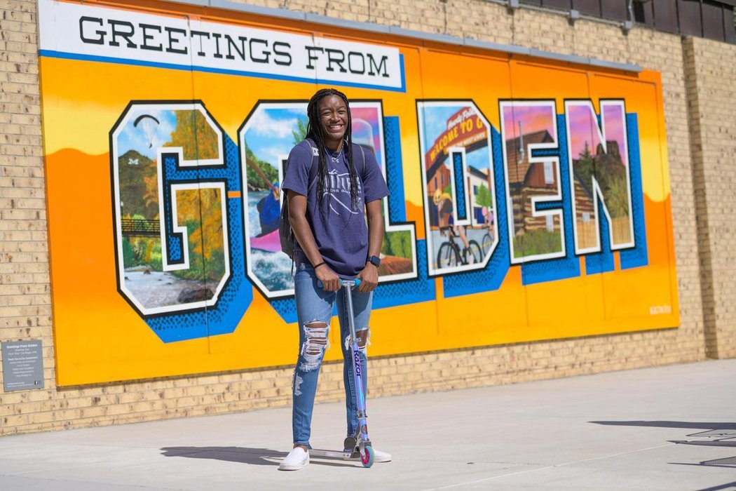 Mines student with a scooter in front of the Welcome to Golden sign