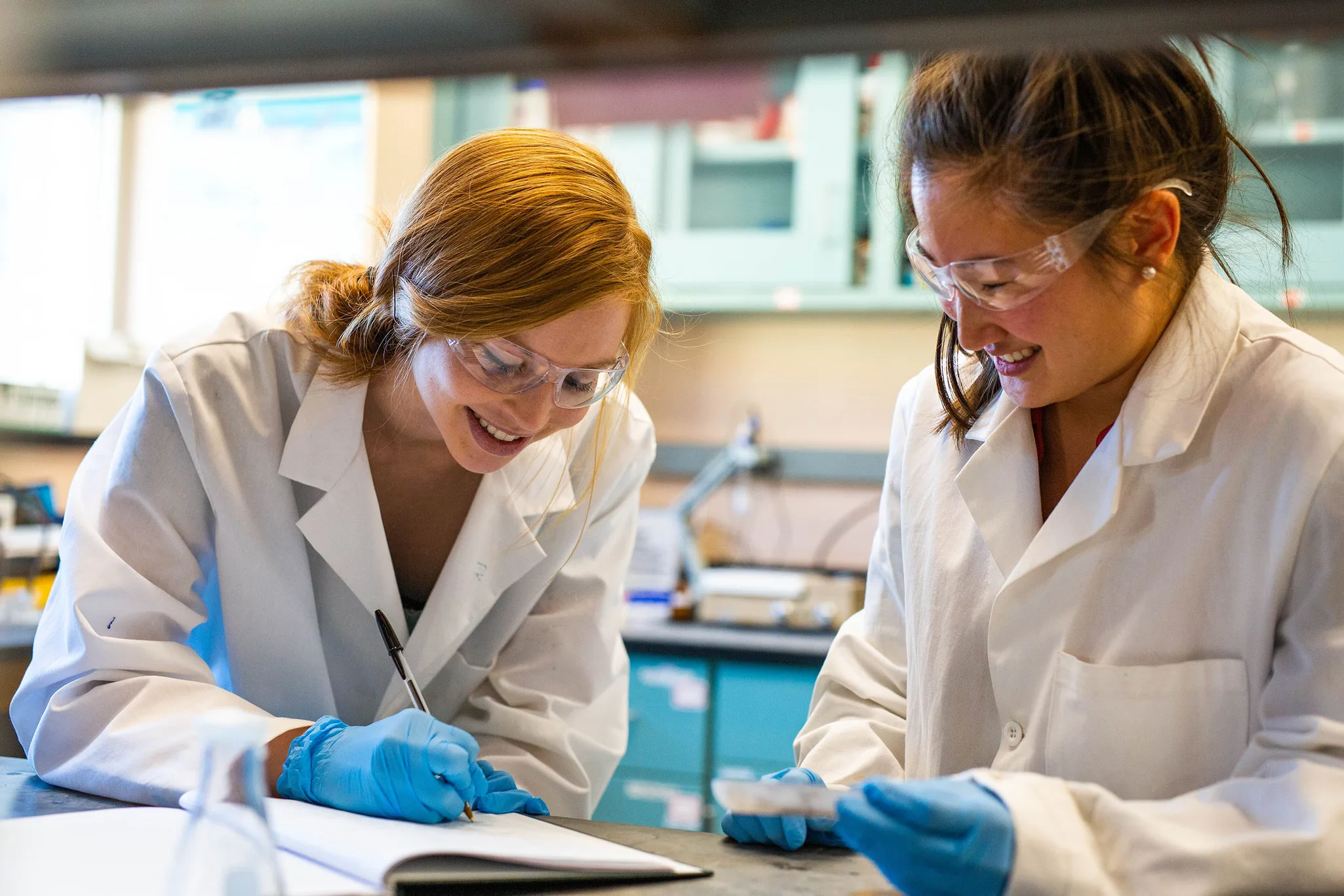 Two female Mines scientists in lab coats and safety goggles work together in a laboratory, one writing notes and the other holding a petri dish.