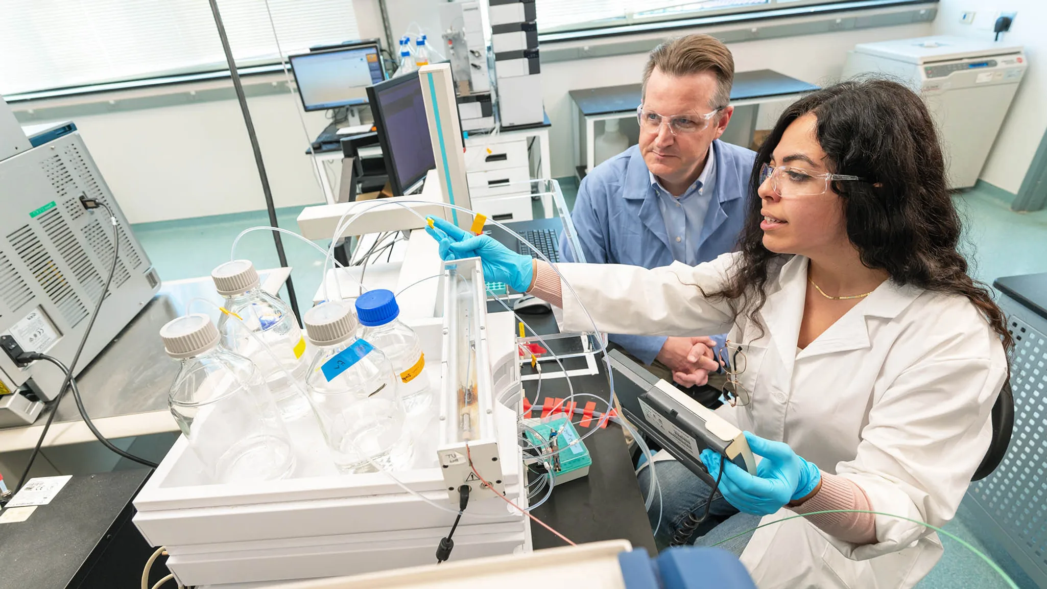 Mines student and professor in a lab coat and gloves operate lab equipment with infusion bottles, tubes, and a computer in a laboratory setting.