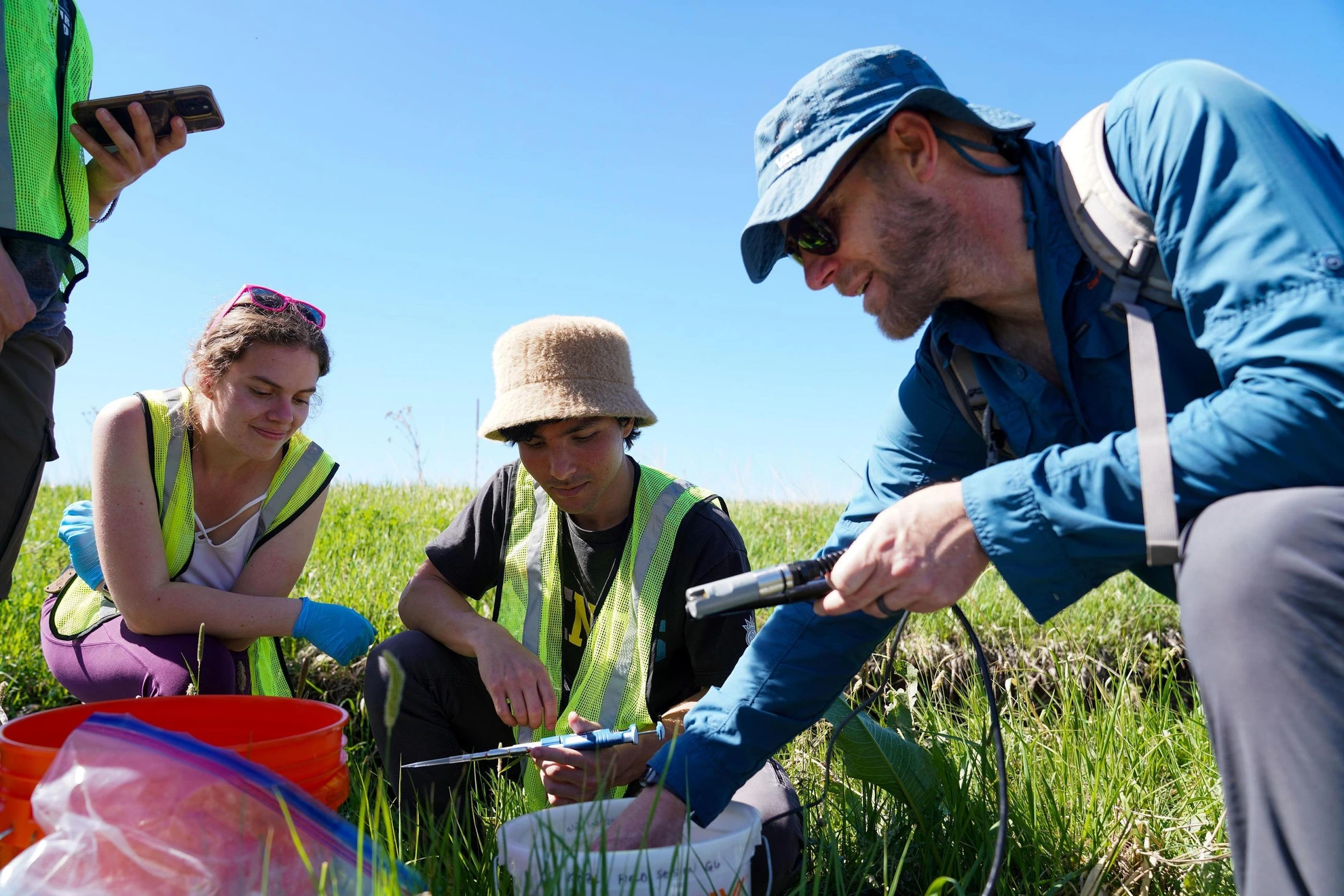 Field researchers in safety vests examine samples and equipment in a grassy field under a clear blue sky.