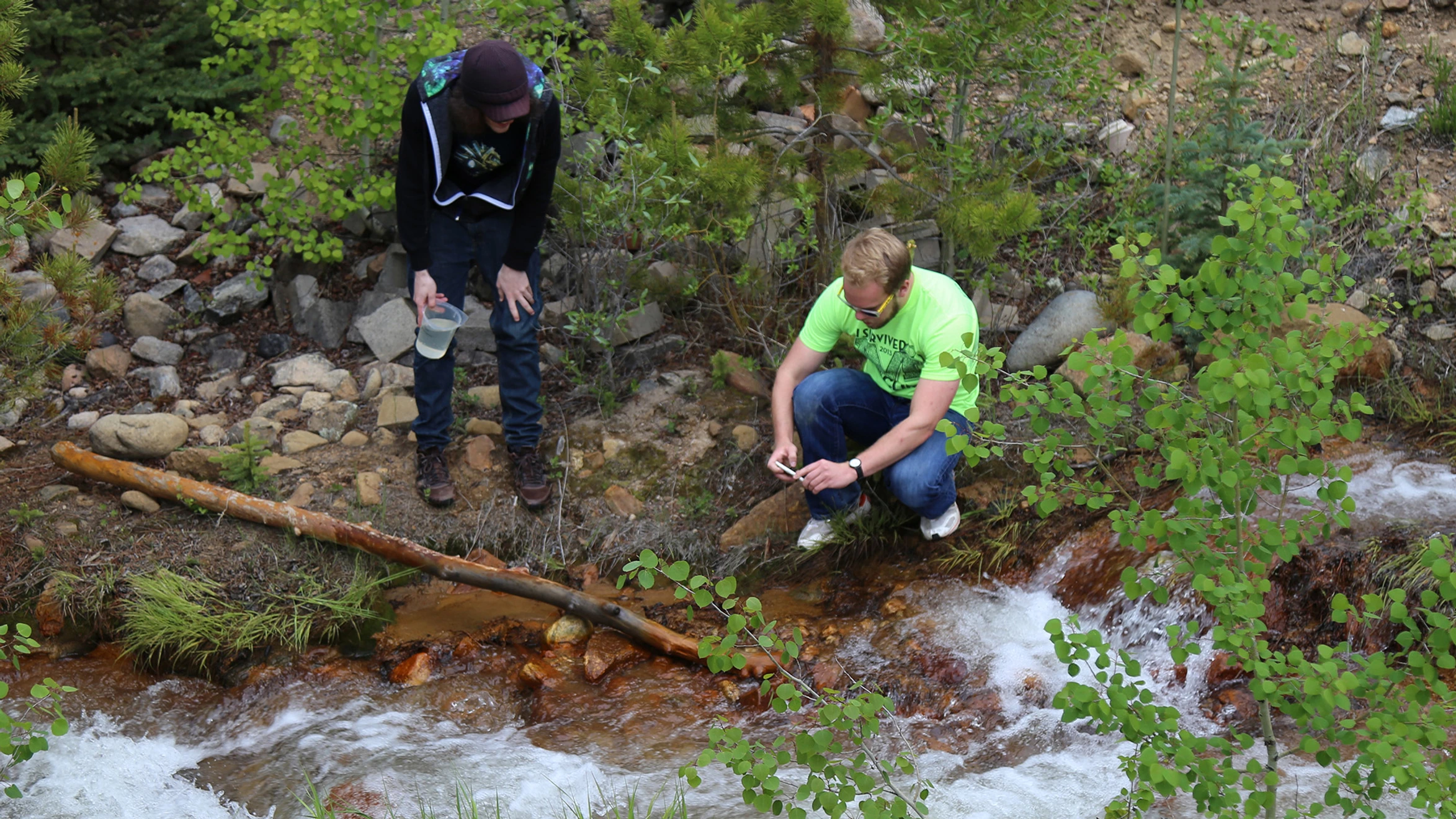 A couple of people standing next to a stream in the woods.