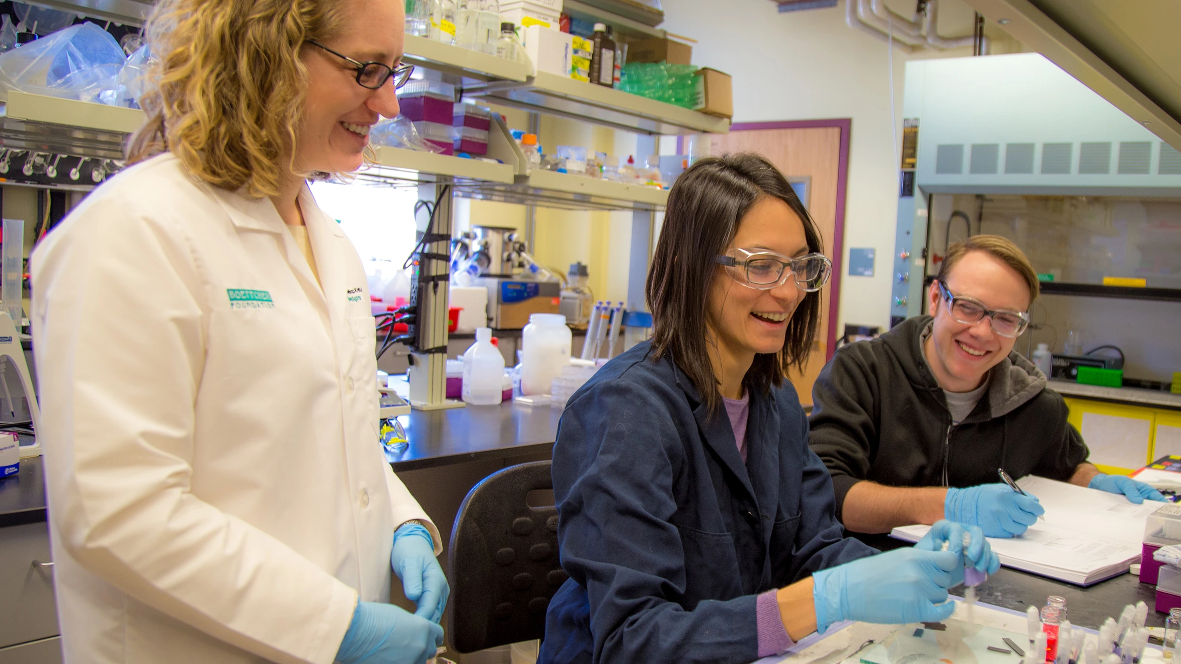 A group of three people in lab coats and gloves working in a laboratory.