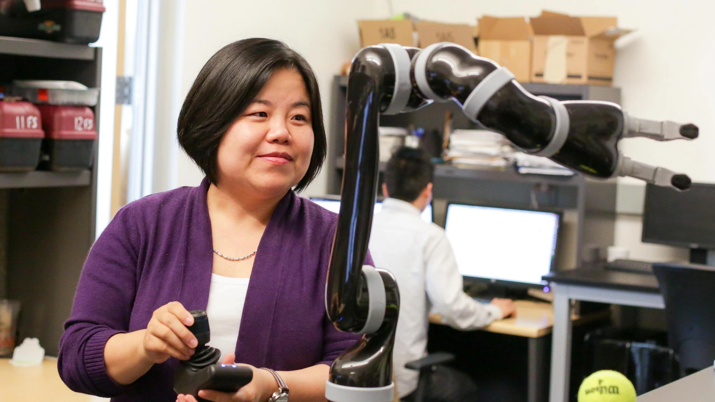 A woman standing in front of a robot holding a tennis ball.
