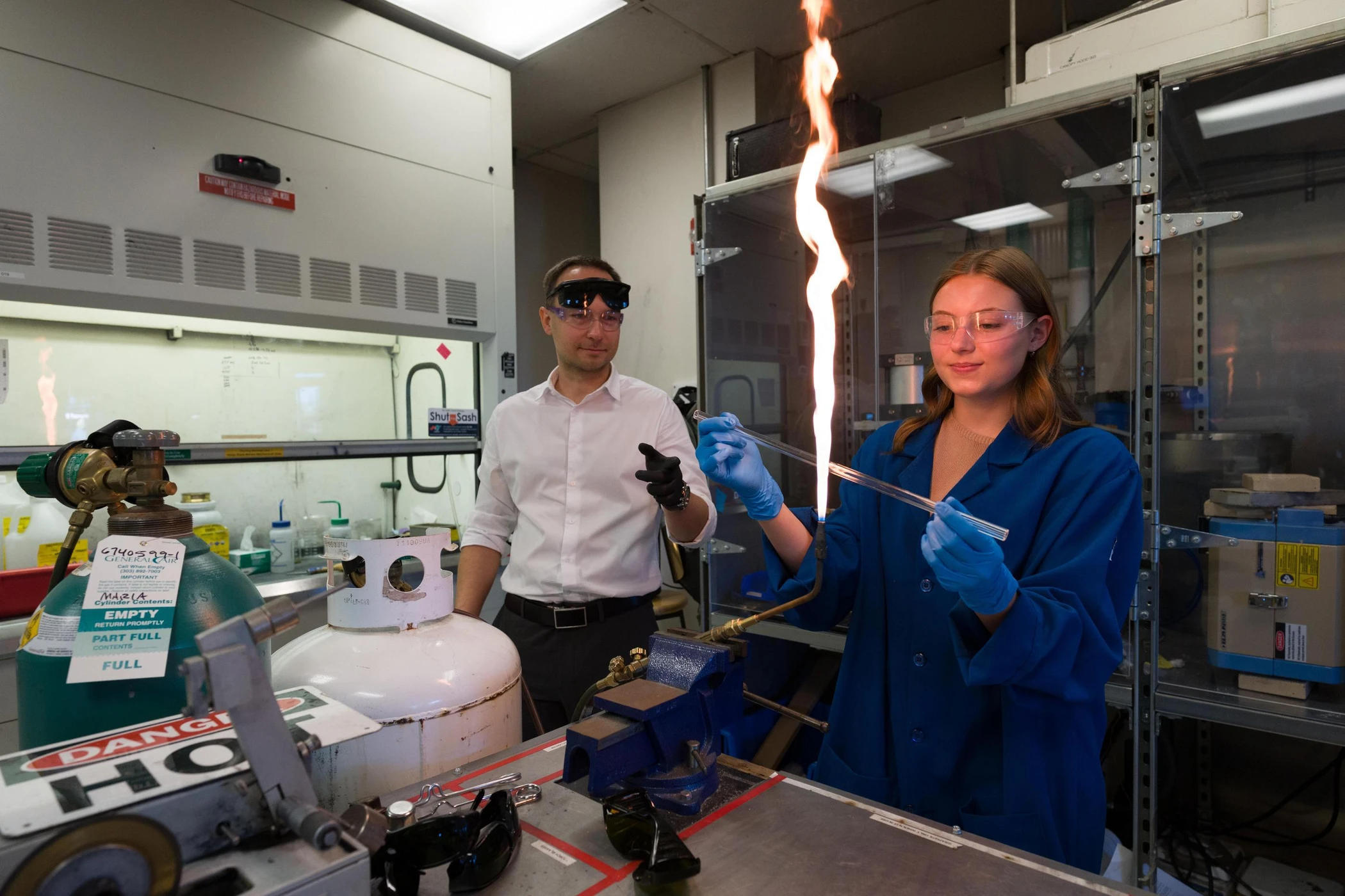 A professor and student in safety glasses and gloves conduct a laboratory experiment with a tall flame and gas cylinders.