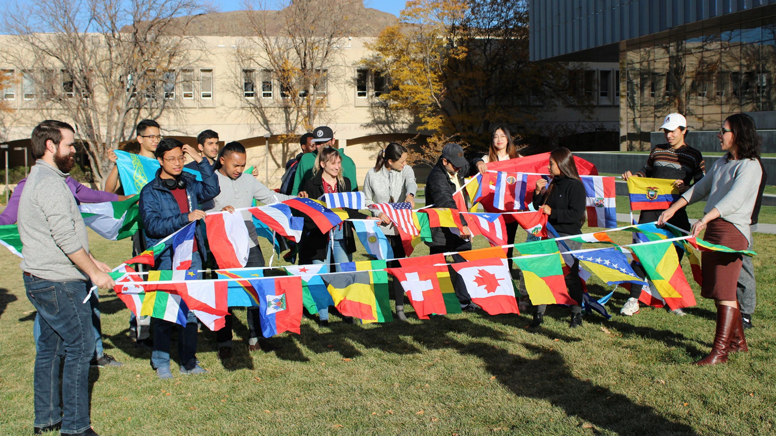 A group of students outdoors holding colorful international flags on strings, standing on grass in front of buildings.