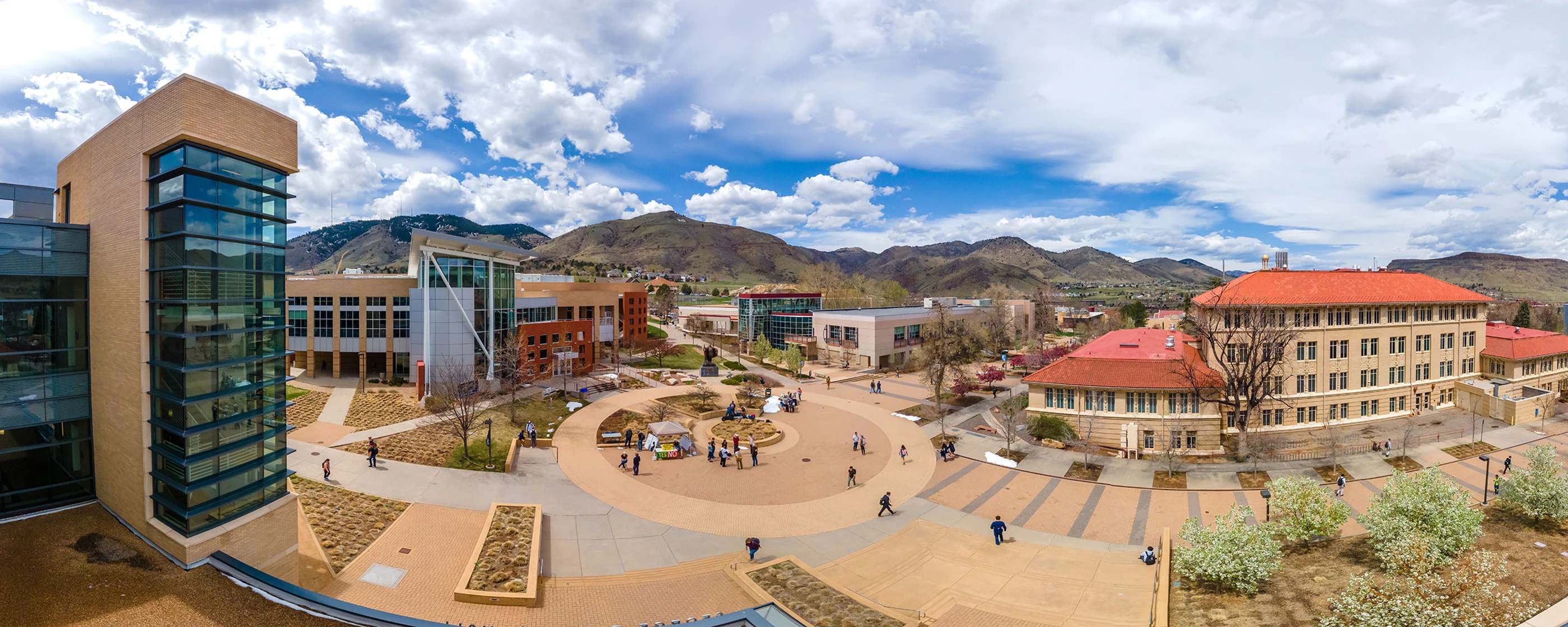 A panoramic view of Mines' campus courtyard with modern and historic buildings, students walking, mountains, and a partly cloudy sky.