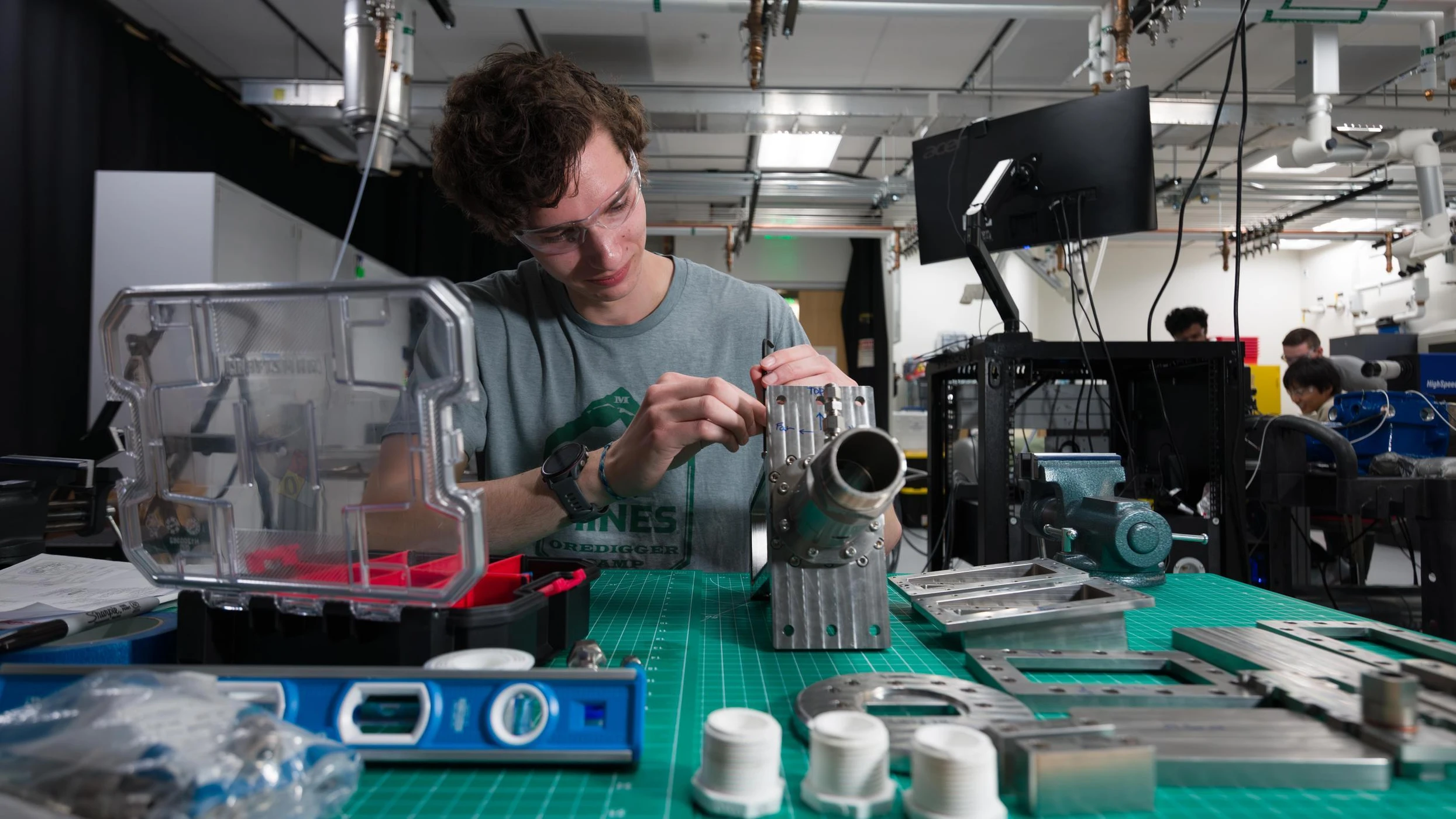 Mines student wearing safety glasses working on a metal mechanical part in a workshop with tools and equipment on the table.