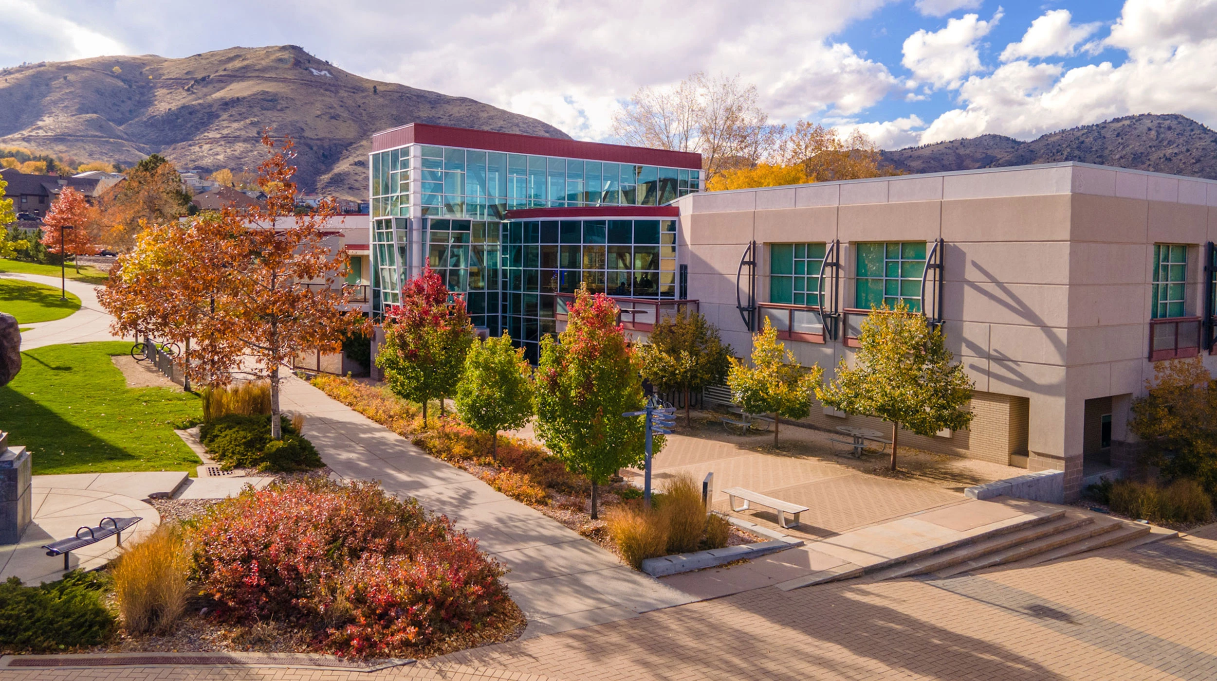 Modern student center campus building with glass windows and autumn-colored trees set against a backdrop of rugged hills under a partly cloudy sky.