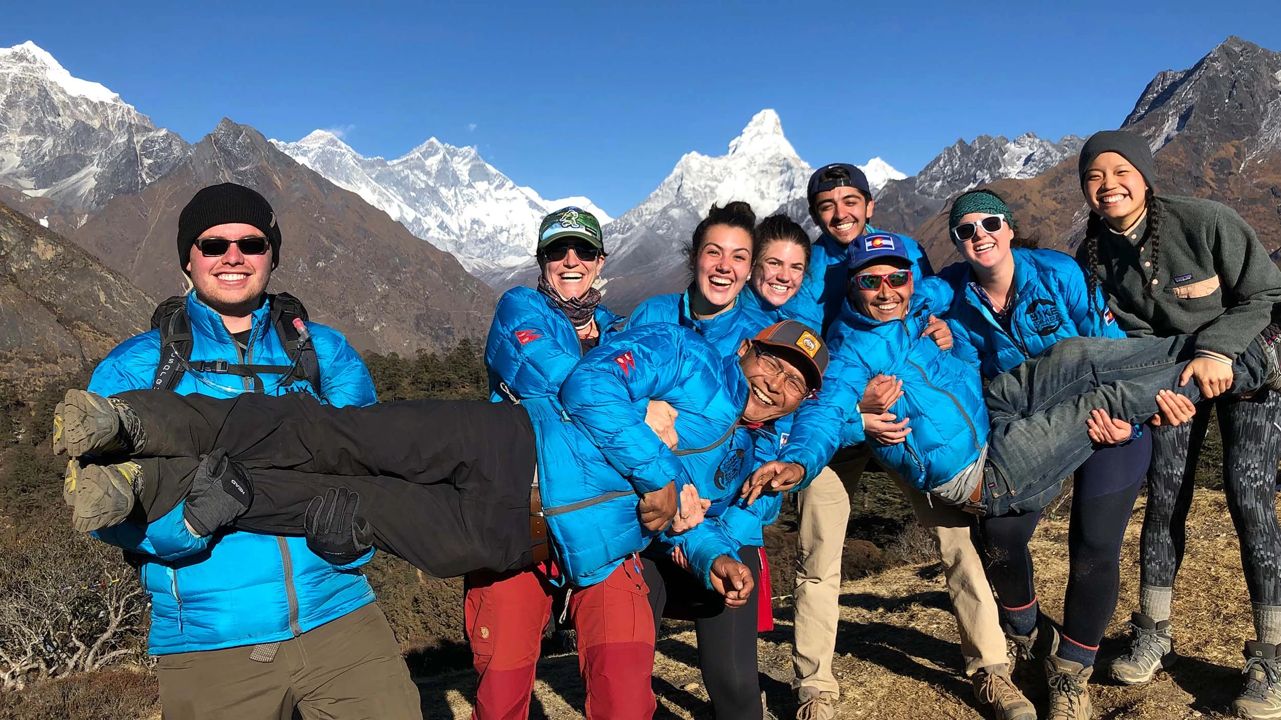 Group of students in blue jackets smiling and holding a person horizontally with snowy mountain peaks in the background.