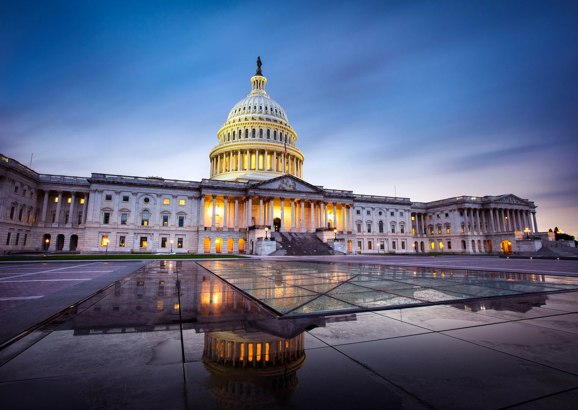 The illuminated United States Capitol building at dusk with its reflection on wet tiled ground in the foreground.