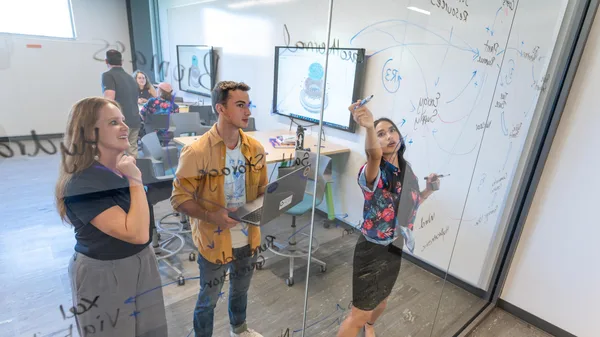 Mines students and a professor work out an organizational operations strategy on a clear glass board in a classroom.