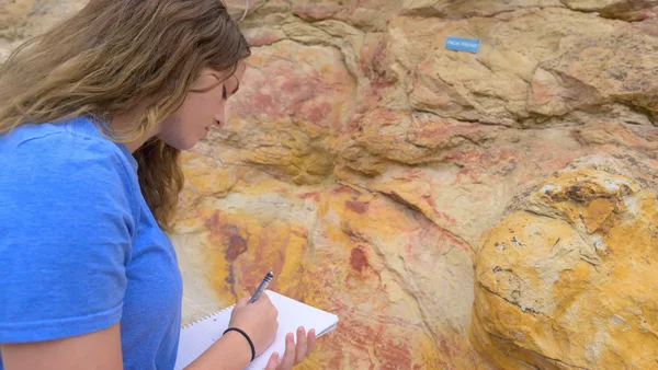 A woman writing in a notebook in front of a rock wall.
