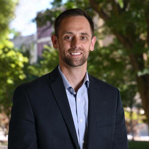 Andrew Lattanner headshot with trees and a university building in the background