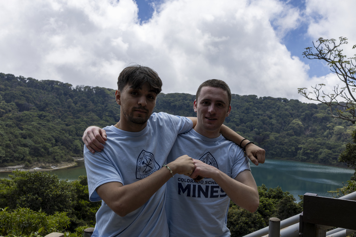 Two male students wearing Mines shirts with their arms around each other fist bumping with a mountainous landscape in the background