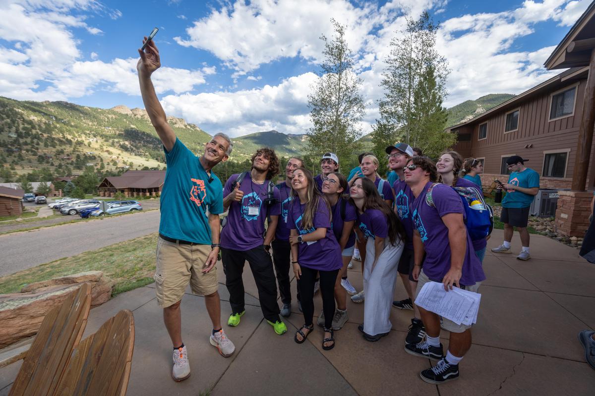 Group of new students in matching t-shirts taking a selfie with President Paul Johnson outdoors near cabins with mountains and blue sky in the background.