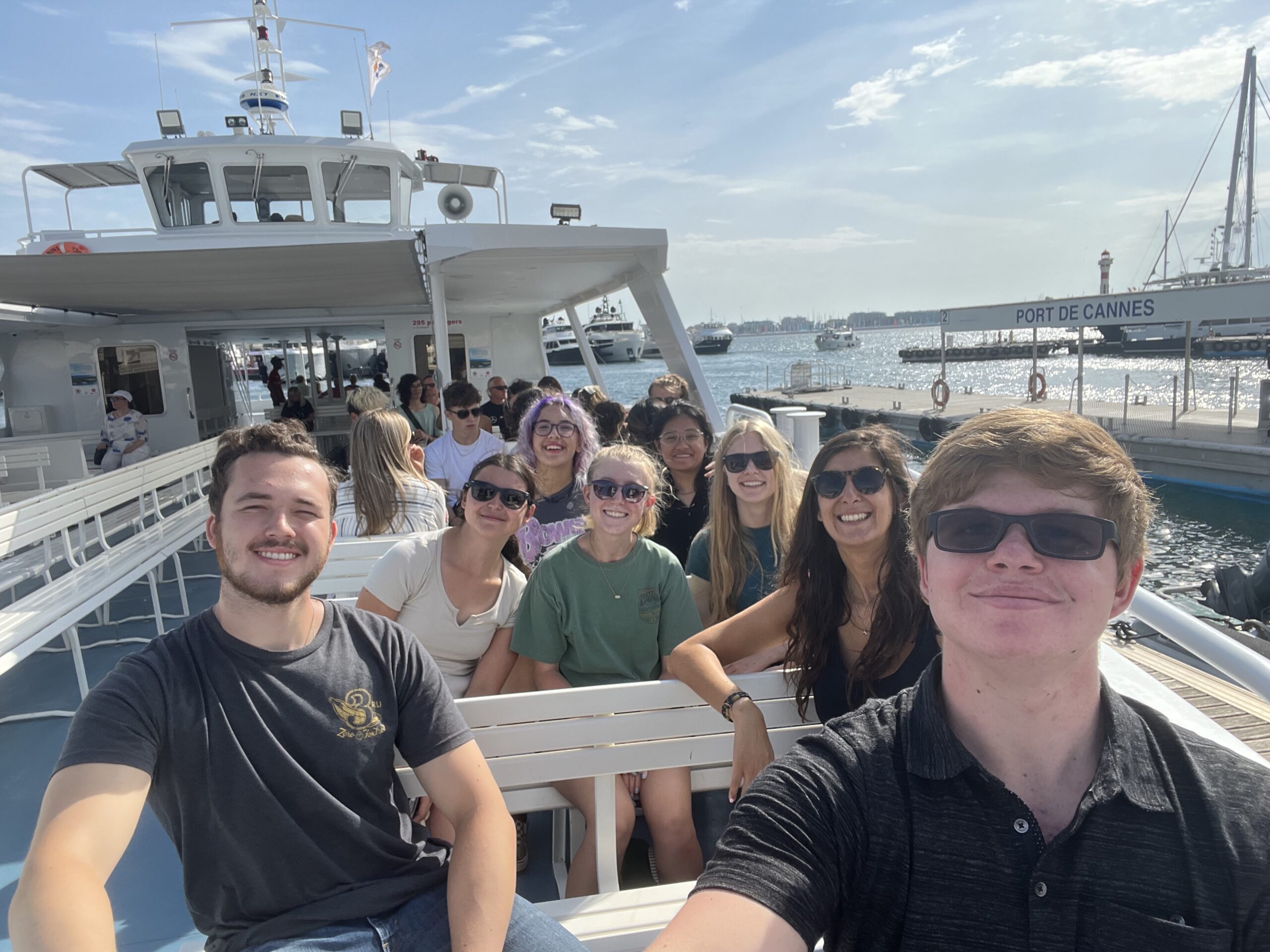 Group of smiling Mines students sitting on a boat at Port de Cannes, with sunny sky and harbor in the background.