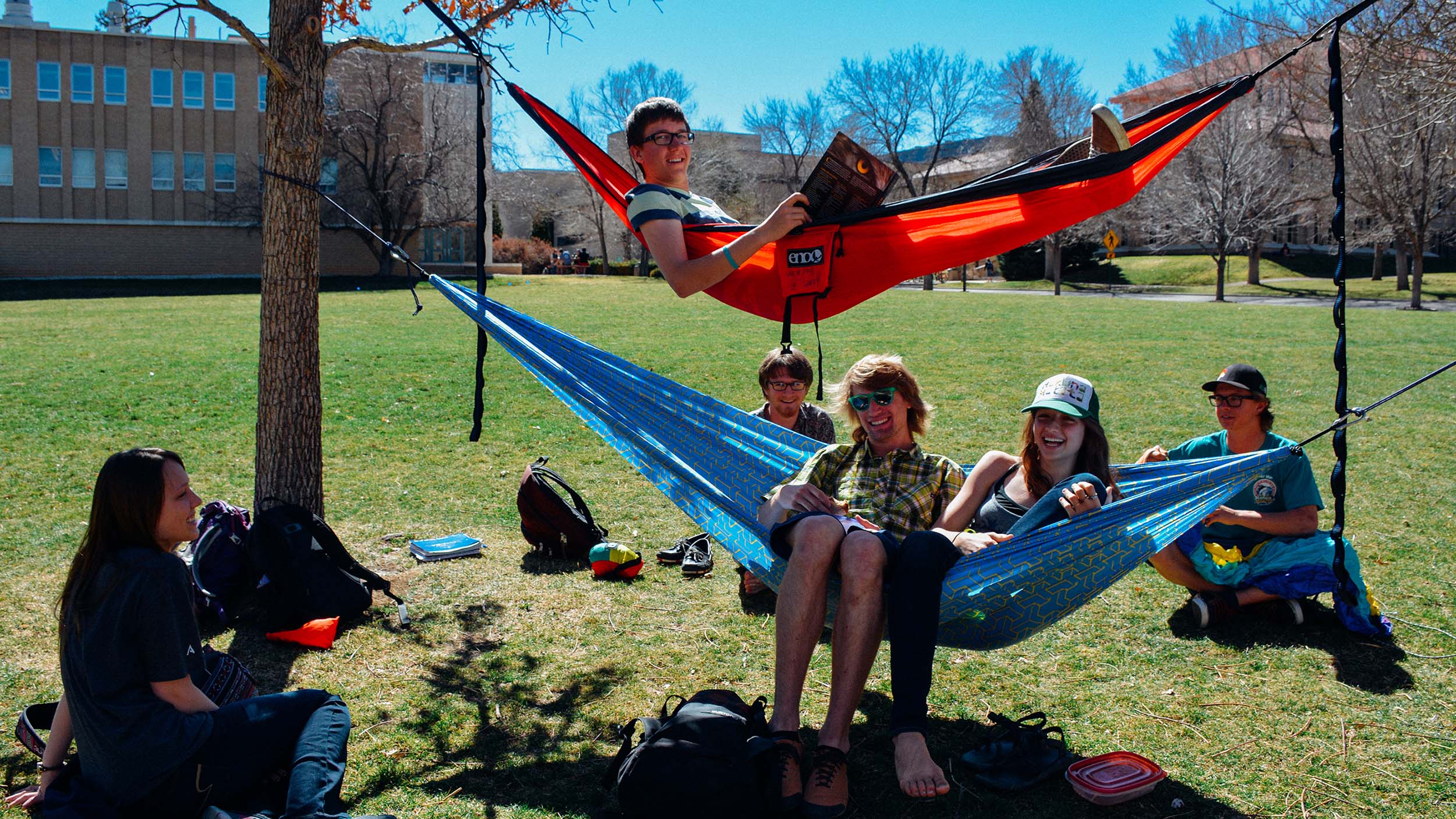Group of six Mines students enjoying a sunny day on a grassy campus, two in hammocks and the rest sitting around them, smiling and chatting.