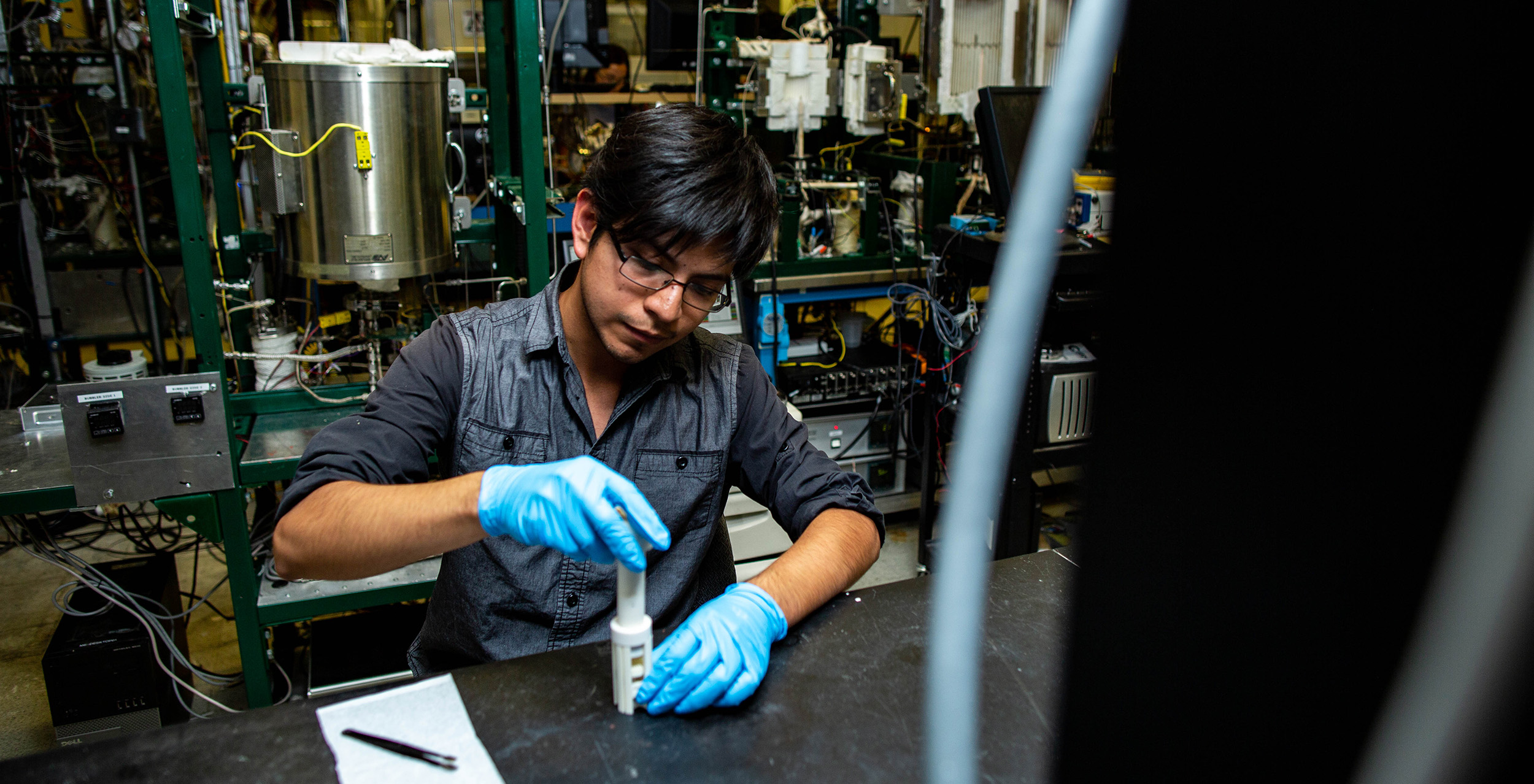 Mines student wearing blue gloves operates a white device on a black table in a lab filled with electronic equipment.
