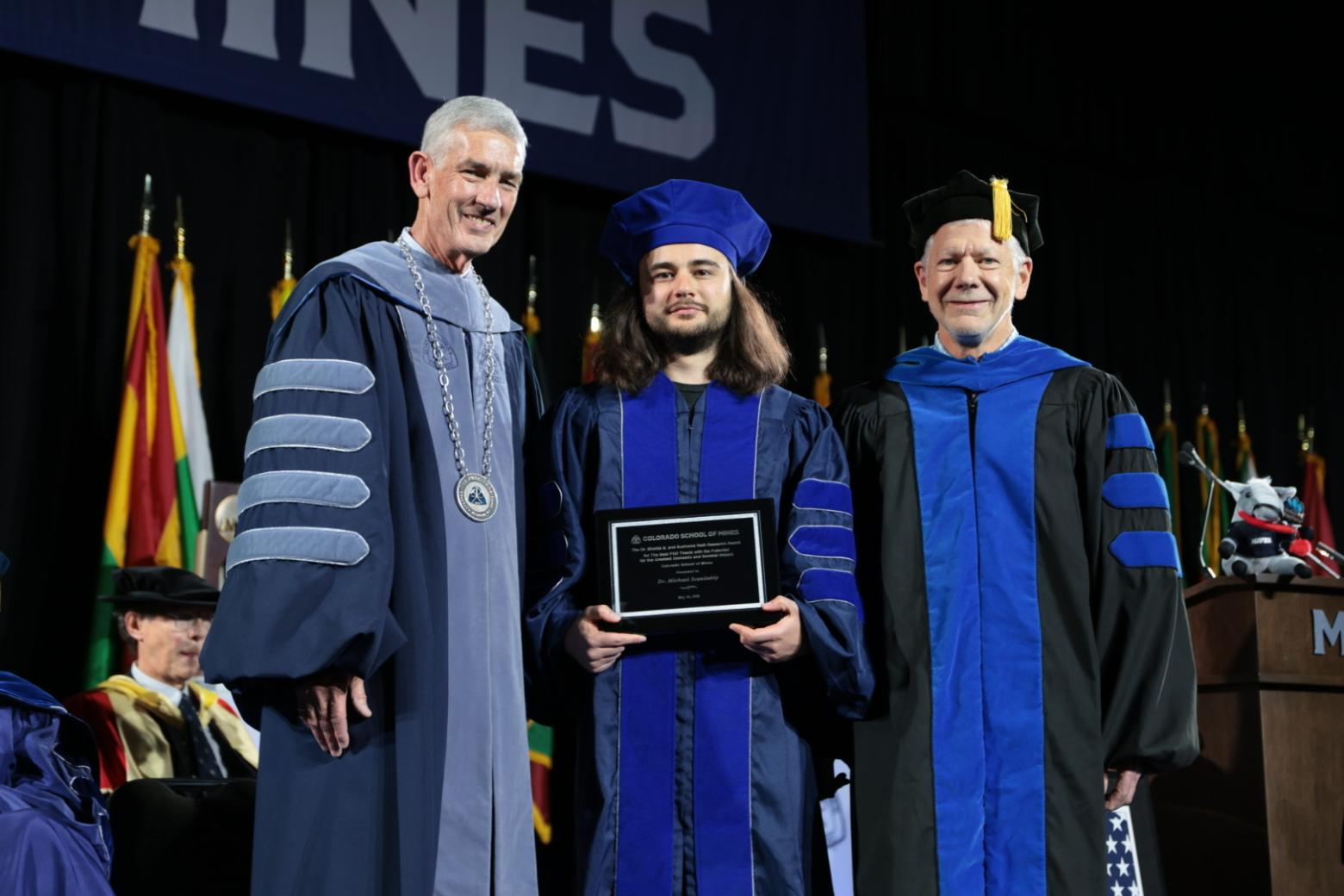 three men in graduation regalia pose together with plaque in the center