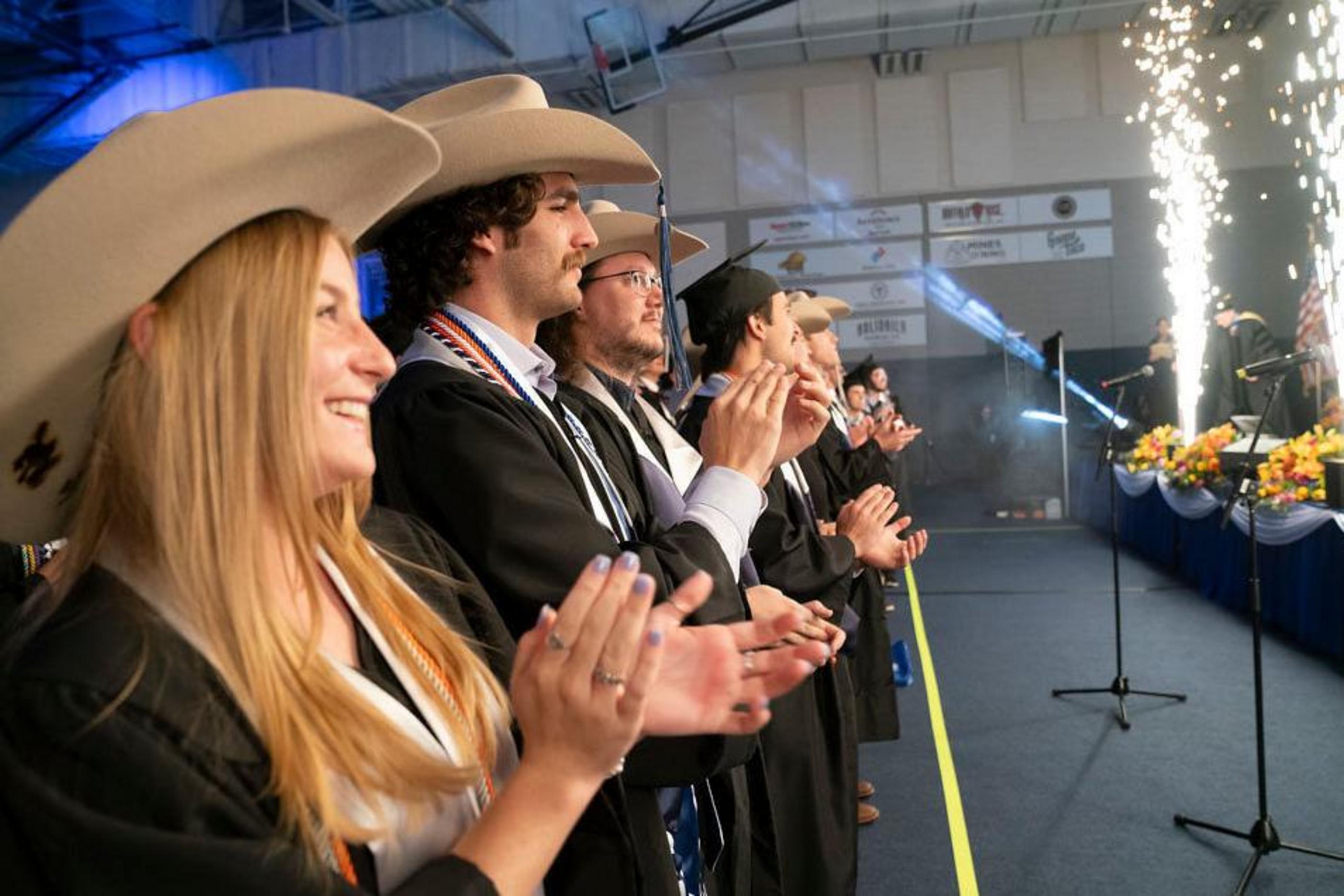 A row of Mines graduates in regalia and Mines Hats applaud during Commencement
