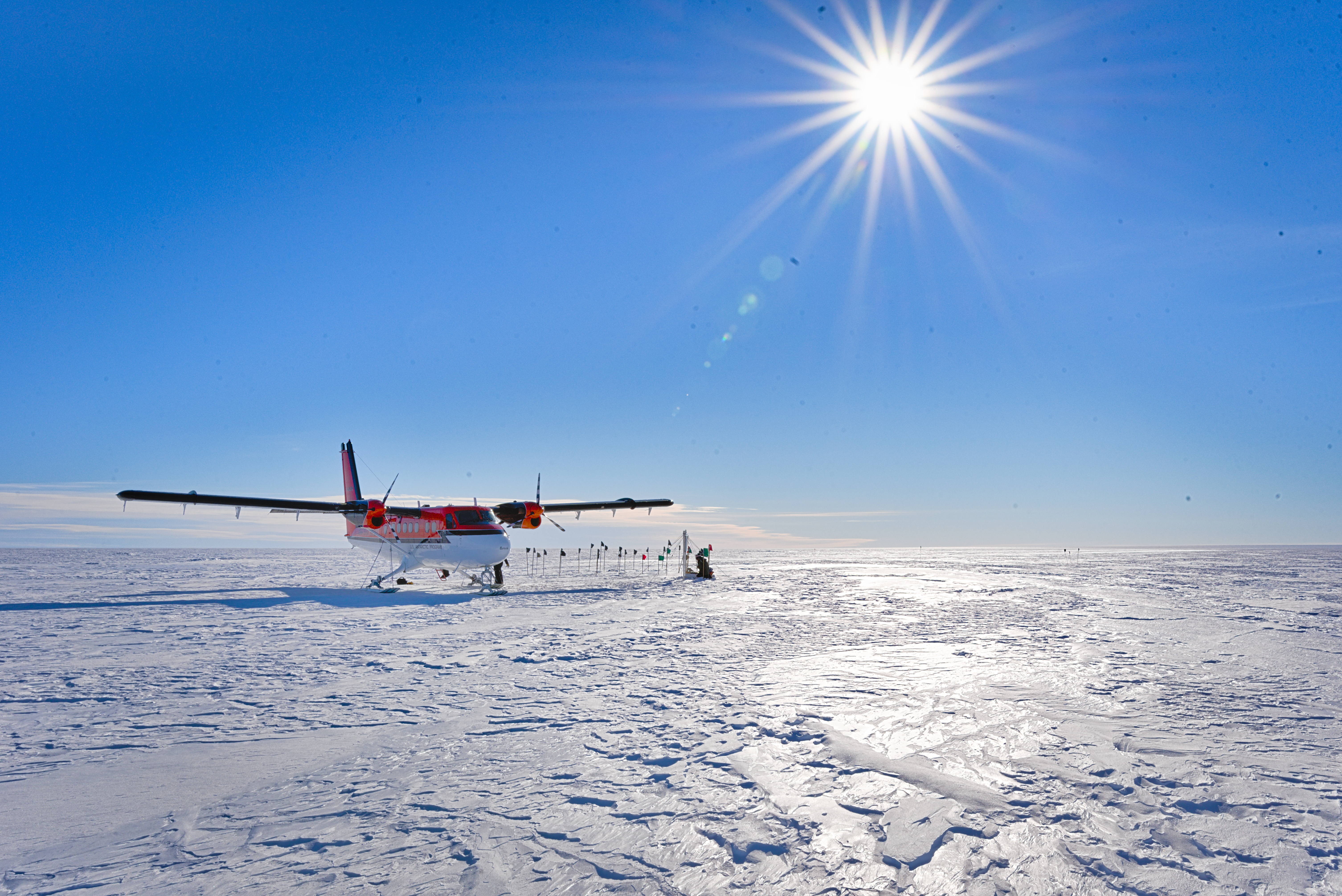 Research plane sits on Antarctic ice with bright blue sky and sun in background