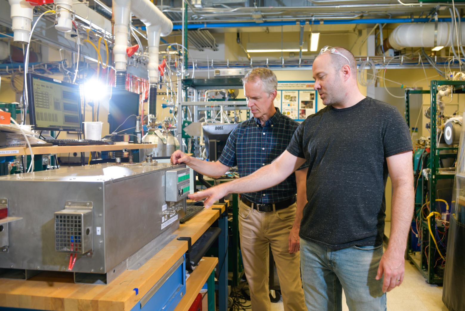 Two men examine and operate a large industrial machine in a lab filled with equipment and monitors.