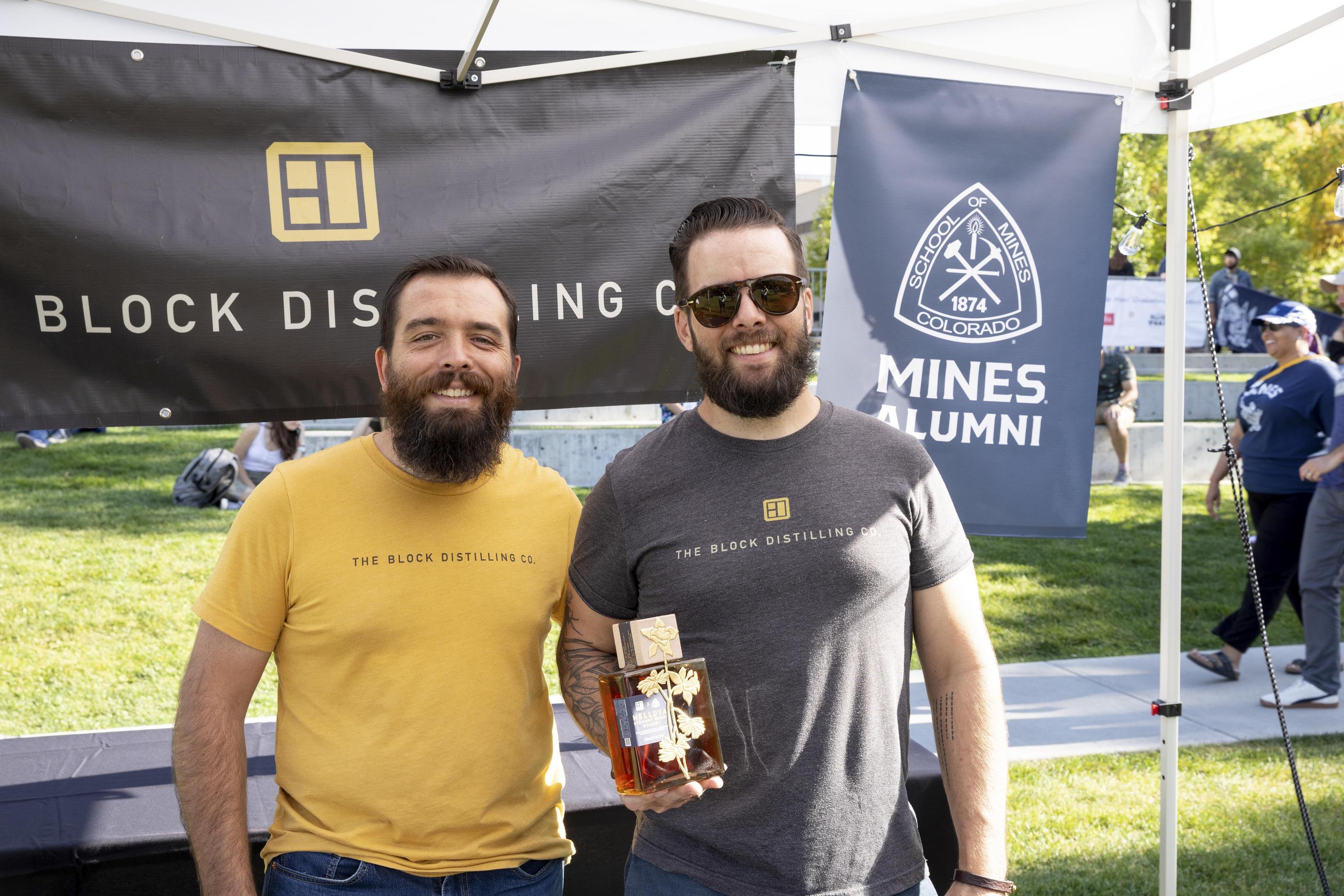 Kameron Weaver '12 and Kraig Weaver '10, co-founders of The Block Distilling Co., smile together at the 2024 Homecoming Blaster's Bash, with Kraig holding a bottle of Helluva Whiskey, the distillery's collaboration with Colorado School of Mines.