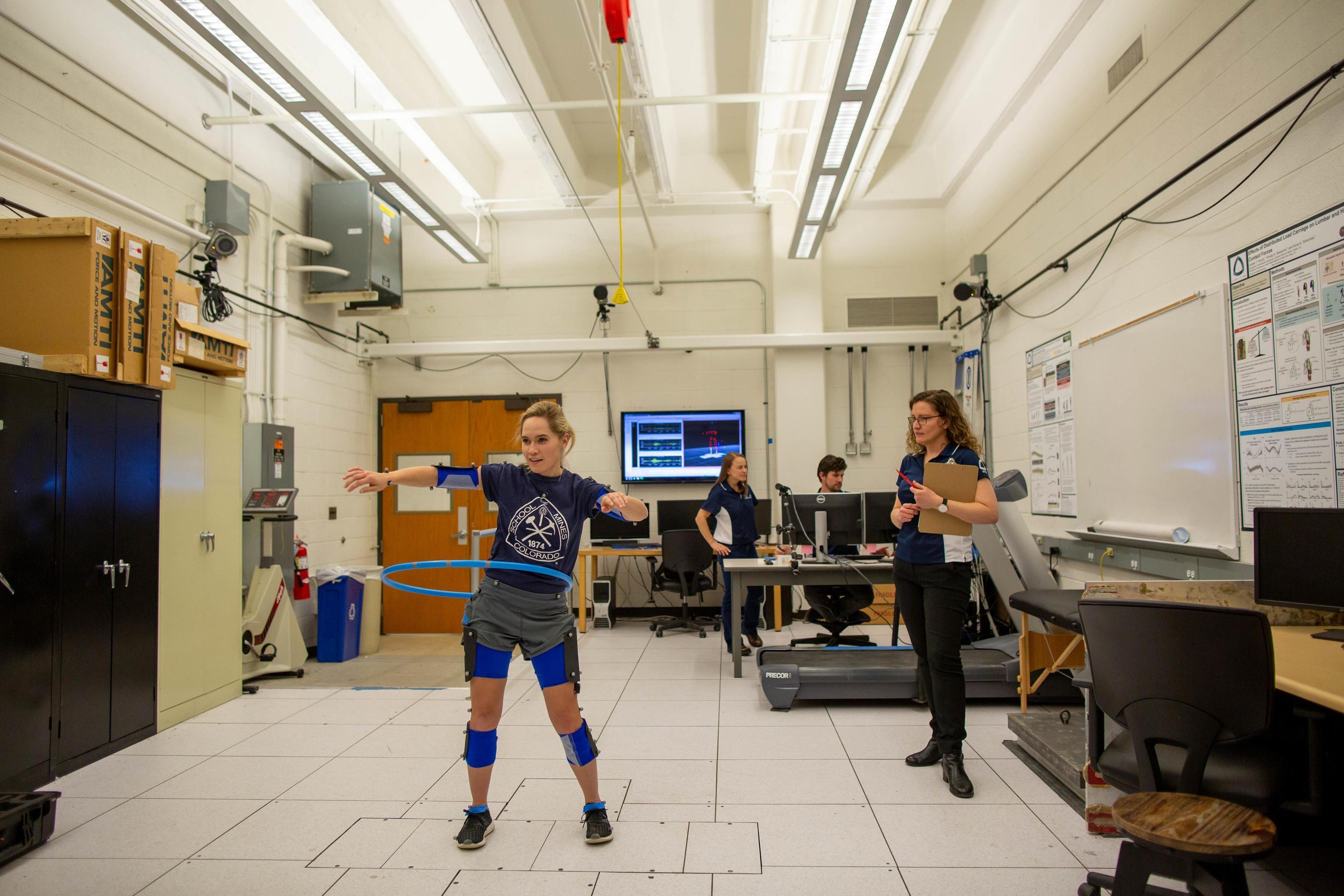 A student wearing biomechanical sensors swings a hula hoop as Anne Silverman observes