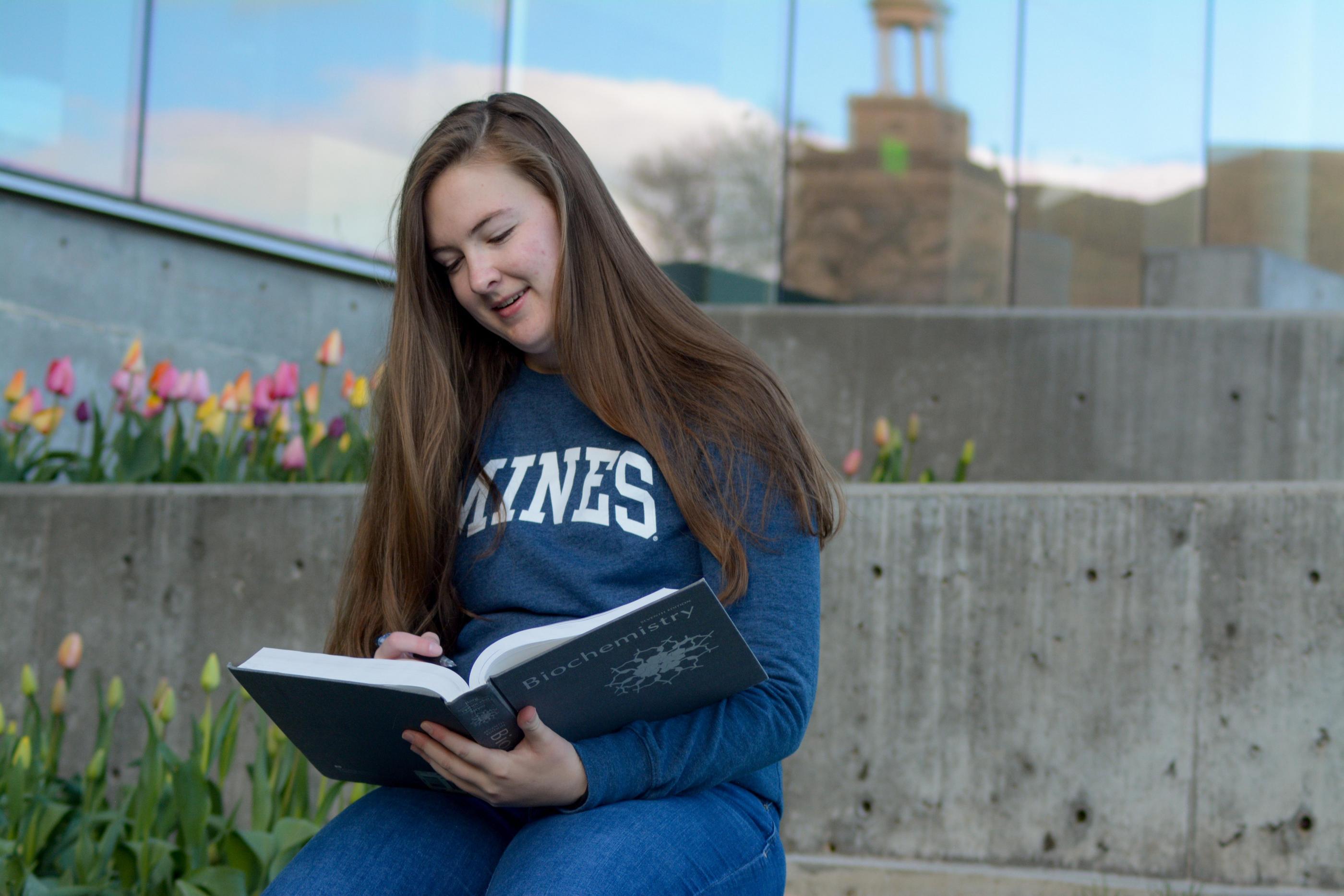 A student in a blue Mines sweatshirt sits outside reading a biochemistry book with reflection of Guggenheim Hall in background