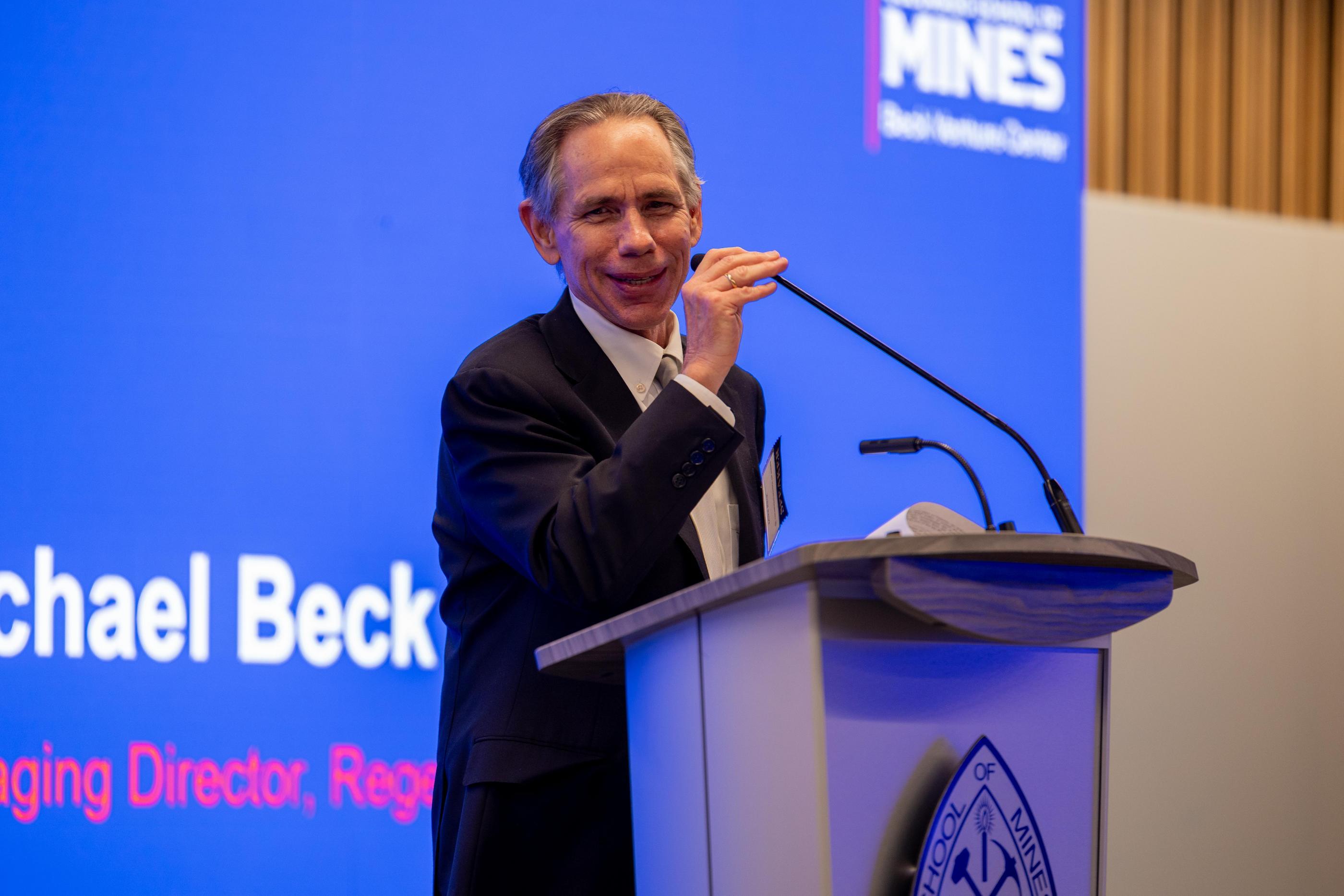 Mike Beck speaks at a podium in front of a blue screen
