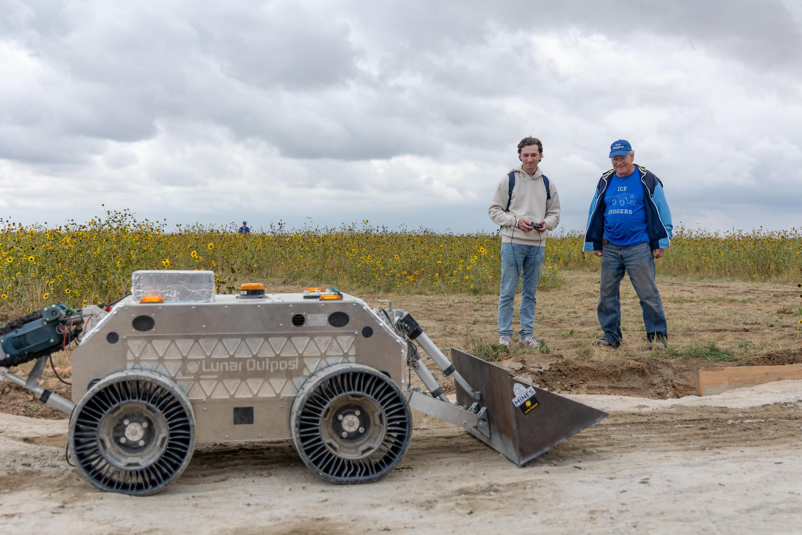 A student and faculty member stand in a sunflower field test driving a lunar rover